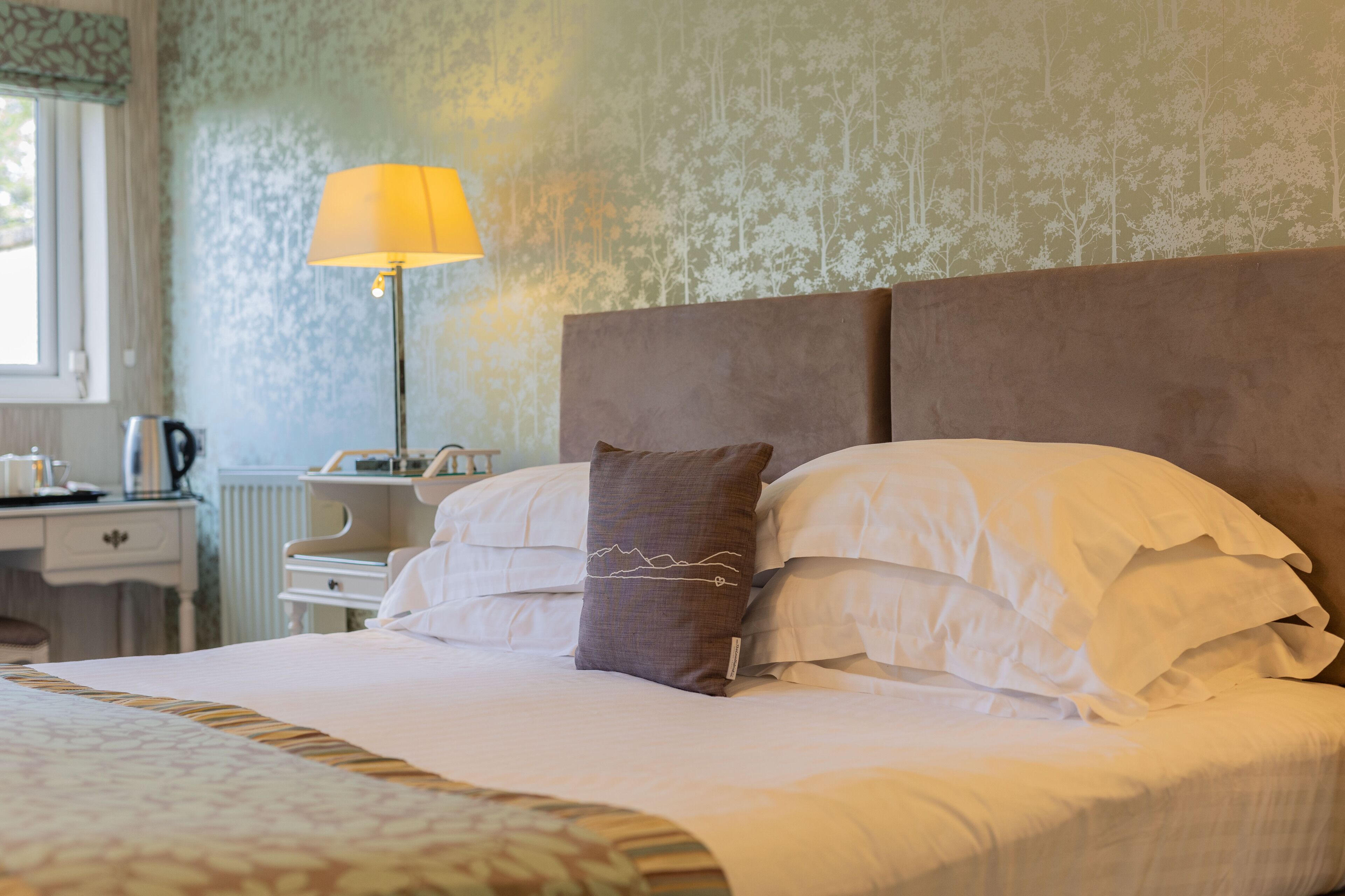Ein ordentlich bezogenes Bett mit weißen Leinen und einem braunen Kopfkissen, ein Nachttisch mit Lampe und eine Wand mit floralem Muster.