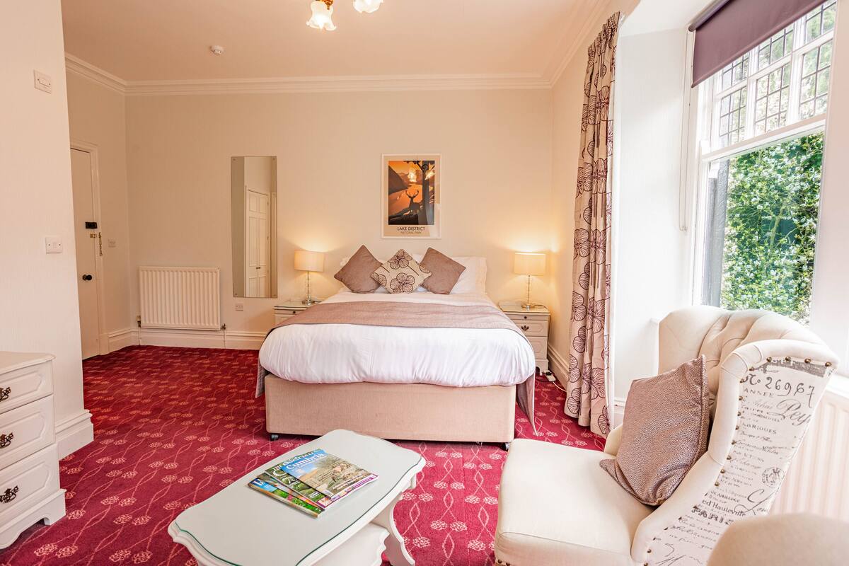 Une chambre d’hôtel avec un grand lit, des tables de chevet avec des lampes, une moquette rouge, une commode blanche et une fenêtre donnant sur un paysage verdoyant.