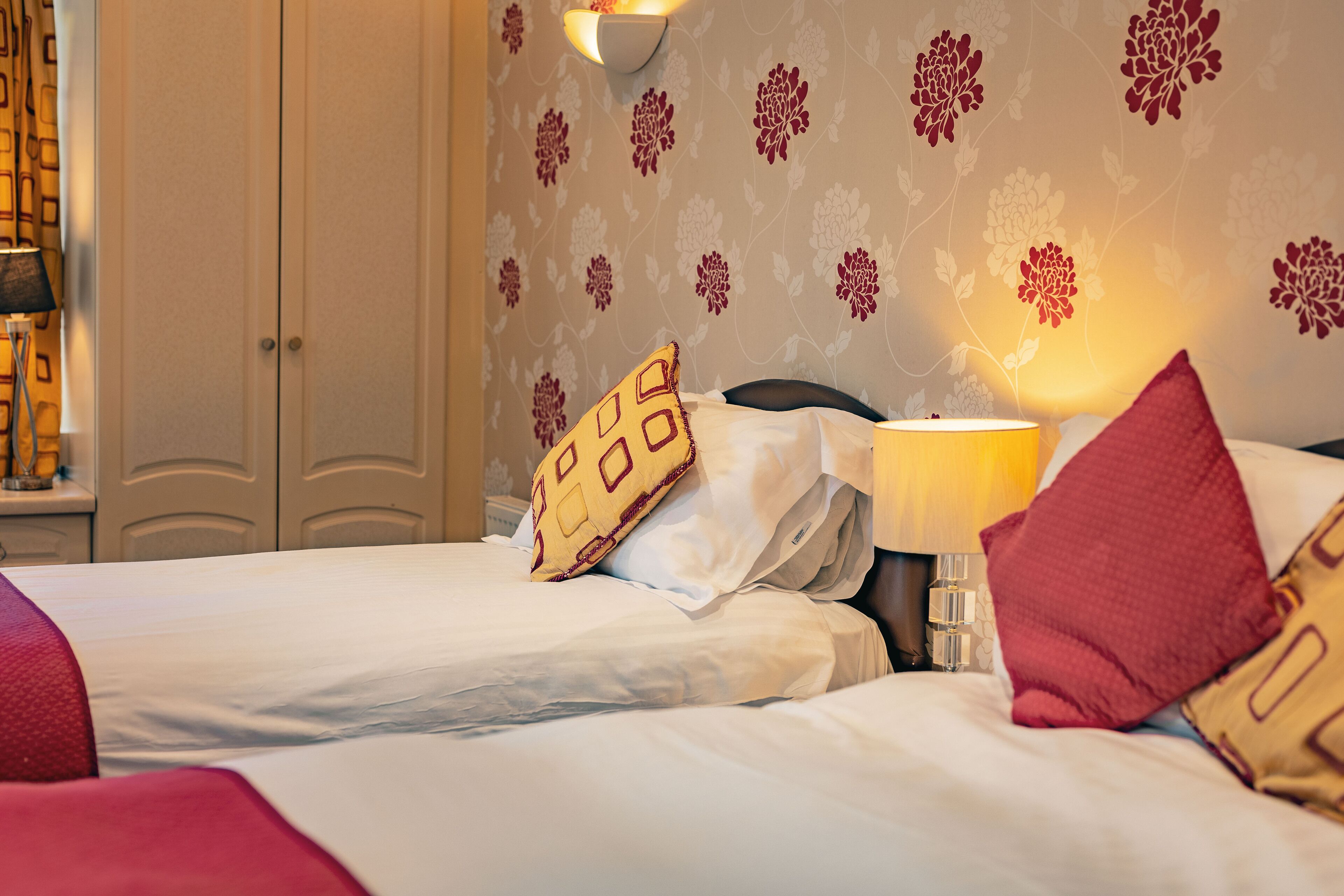 Ein Hotelzimmer mit zwei Betten, einem Nachttisch mit Lampe und blumigem Wandpapier.