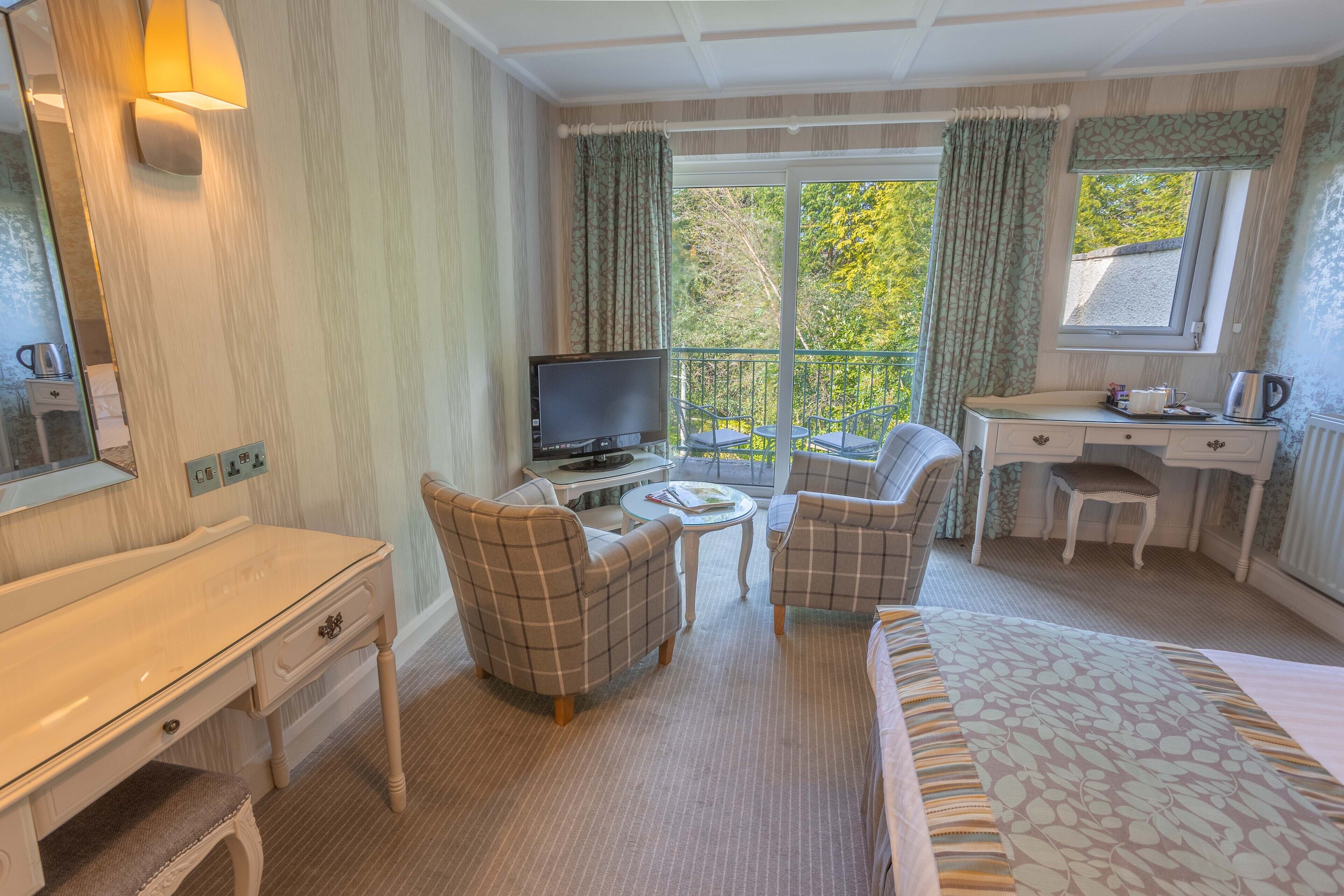 Ein Hotelzimmer mit Bett, Schreibtisch, Fernseher, zwei Sesseln, einem kleinen Tisch und einem Fenster mit Vorhängen.