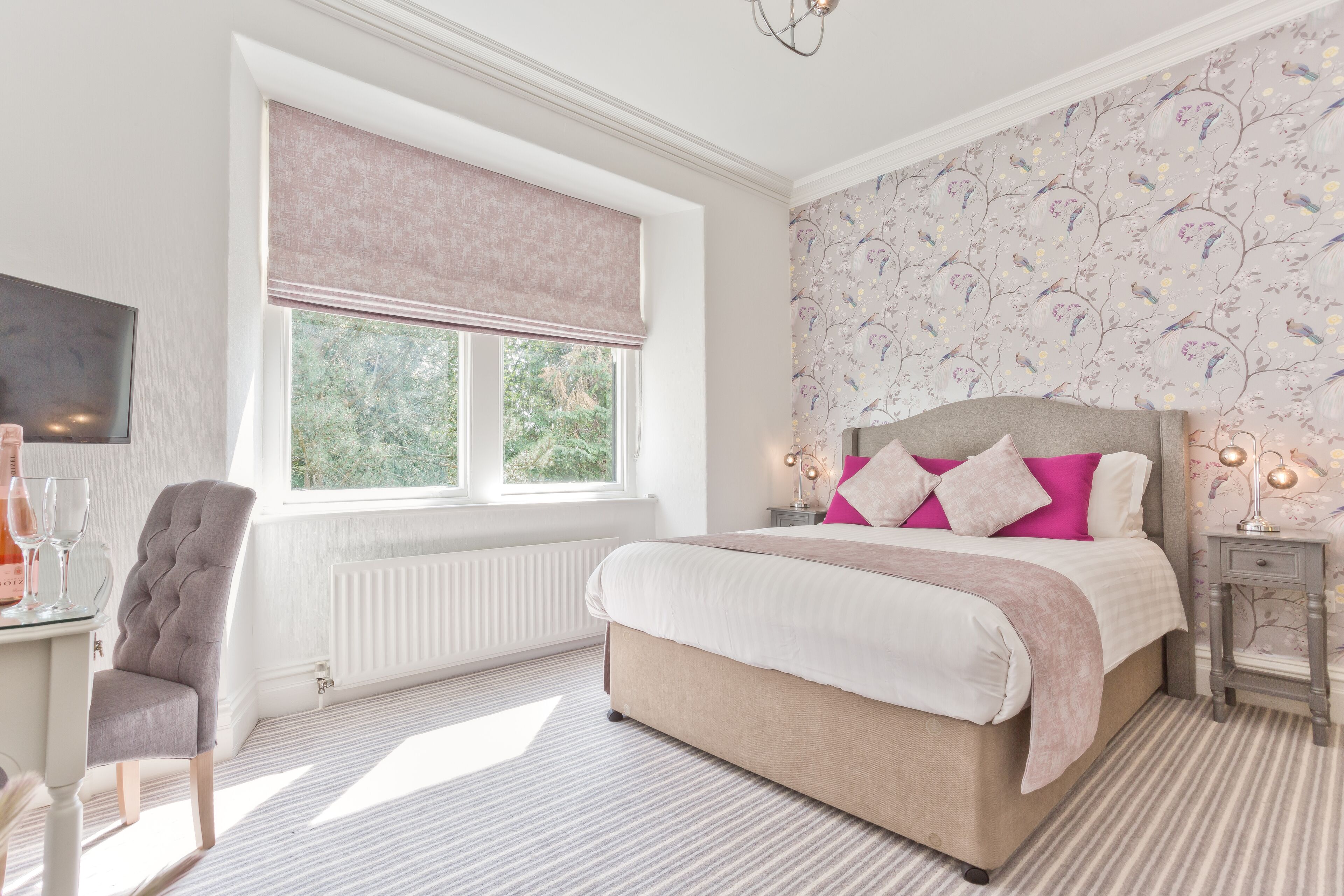Un dormitorio con una cama grande, una ventana con persiana enrollable, una mesita de noche con lámparas y una silla.