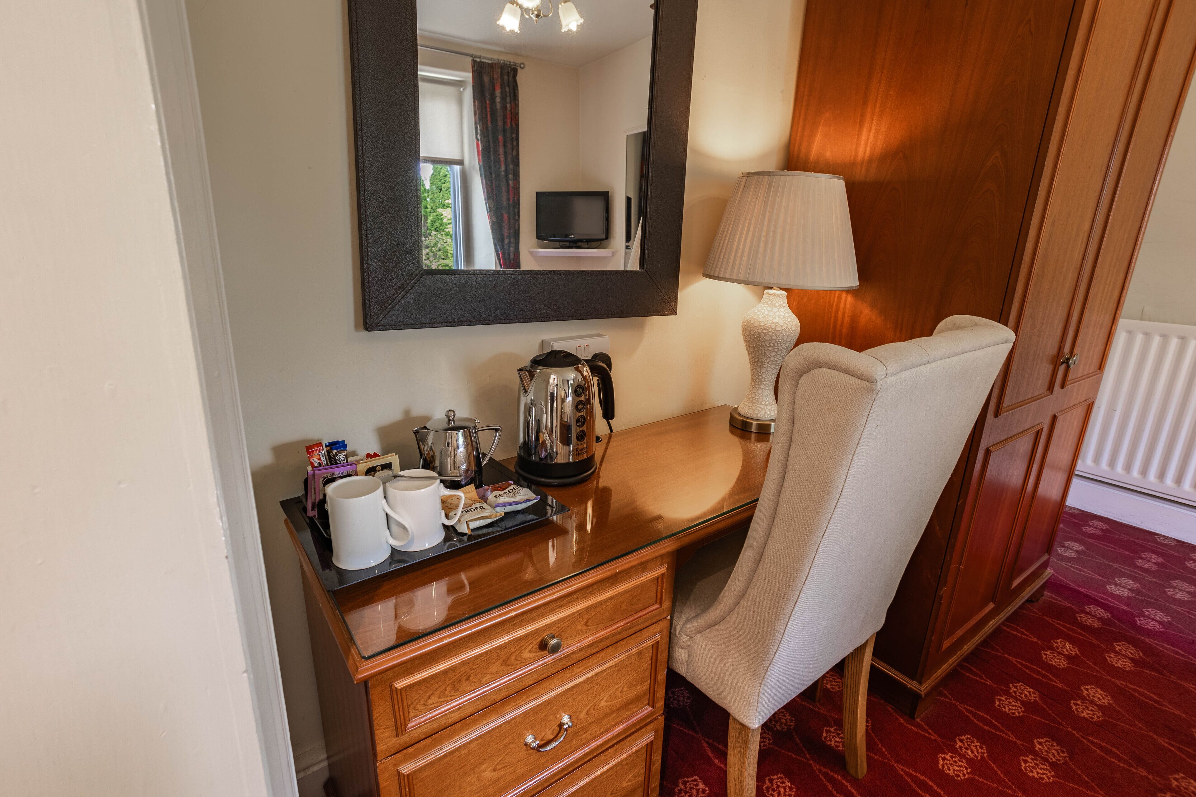 Habitación de hotel con escritorio de madera, una silla, un espejo, televisión, lámpara y radiador.