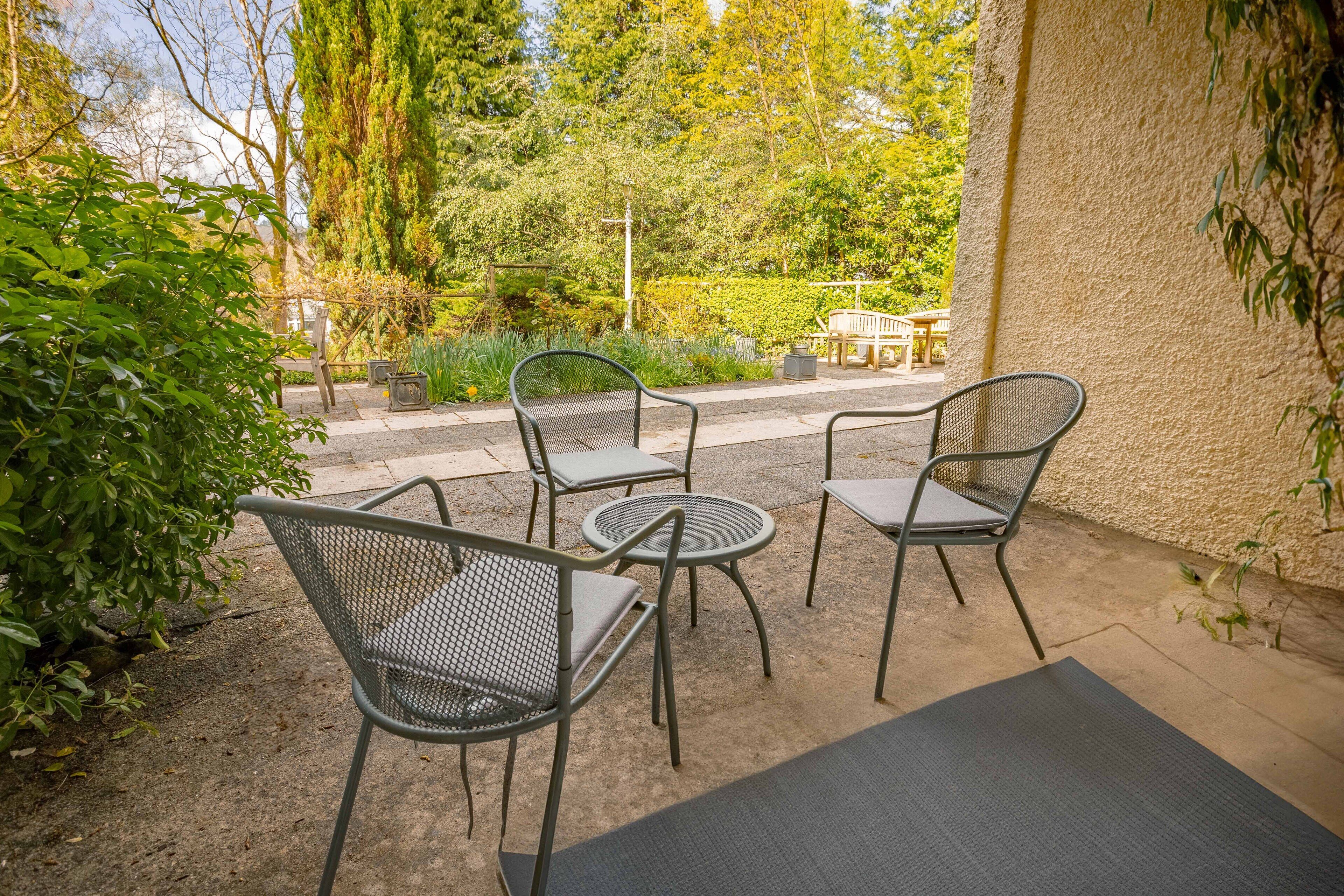 An outdoor patio with metal chairs and tables, surrounded by greenery and a view of the street.