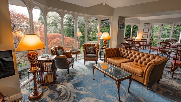 A spacious living room with a large brown leather sofa, two armchairs, a coffee table, and a bookshelf. Large arched windows provide a view of the garden. - The Burn How Garden House Hotel (Windermere)
