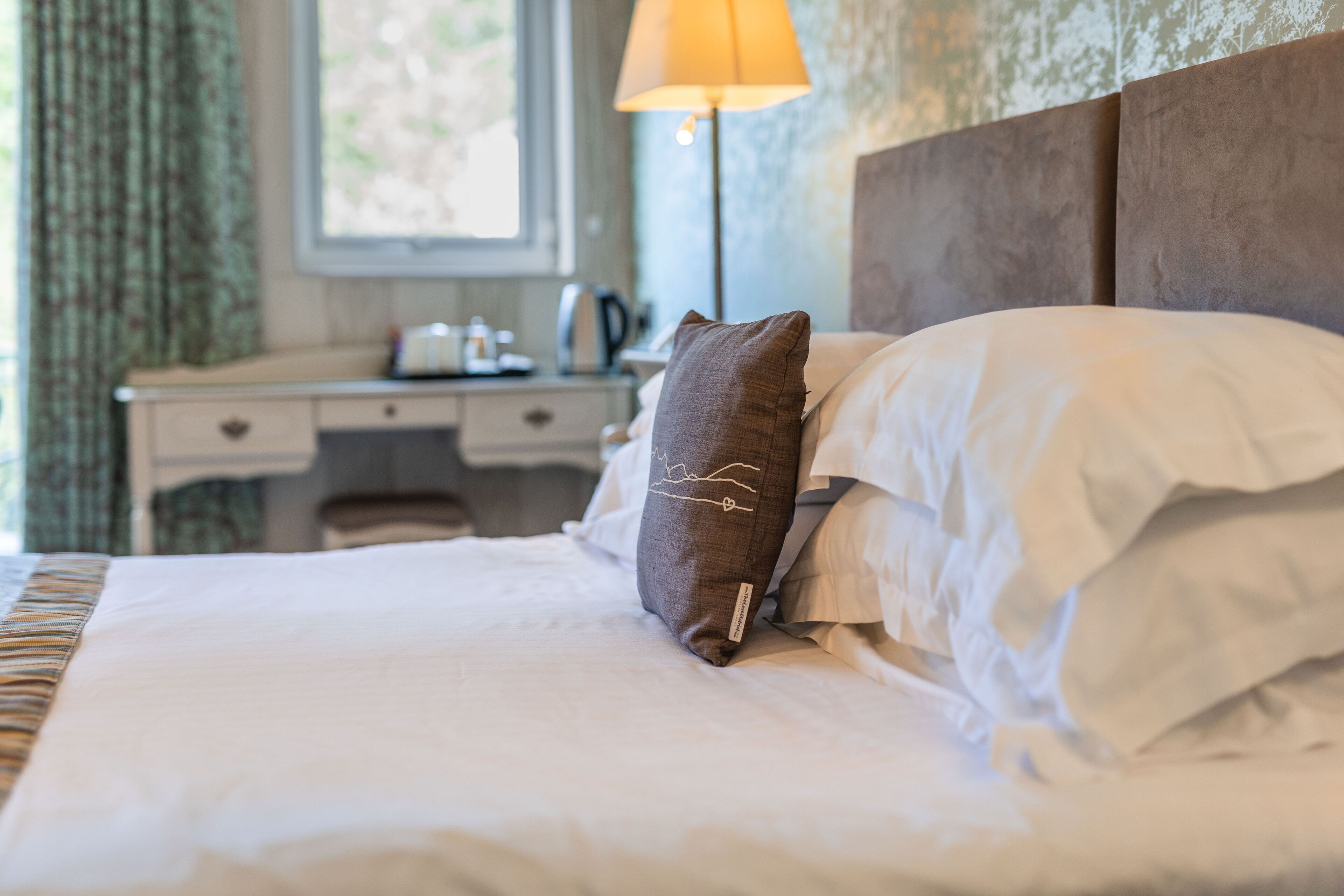A neatly made bed with white linens and a brown pillow, a bedside lamp, and a desk with a kettle.