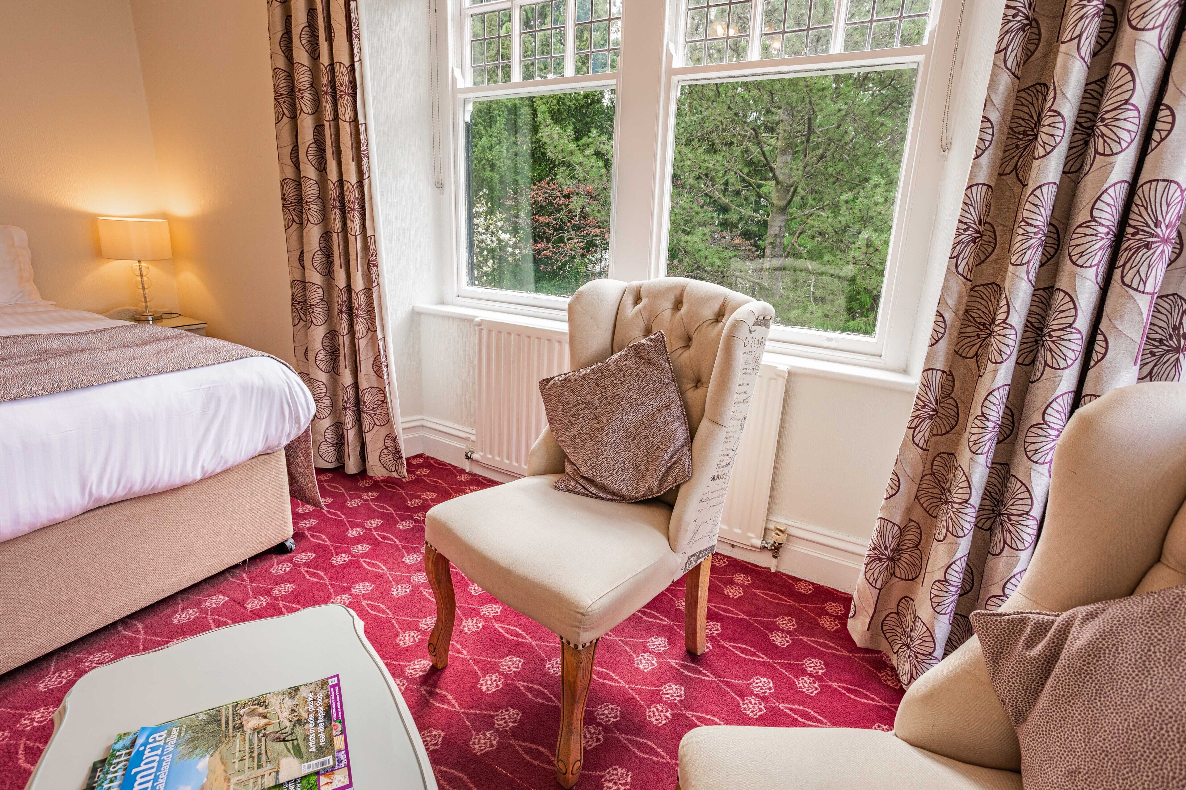 A hotel room with a bed, a chair, a table with a magazine, and a window with curtains.