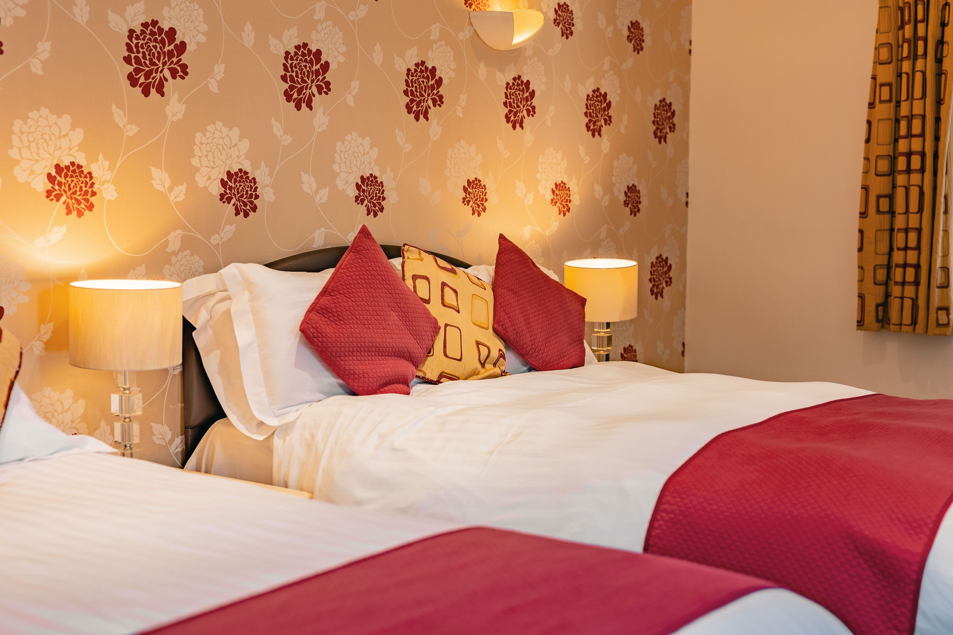 A neatly made bed with red and white bedding, two bedside lamps, and a floral wallpaper pattern.