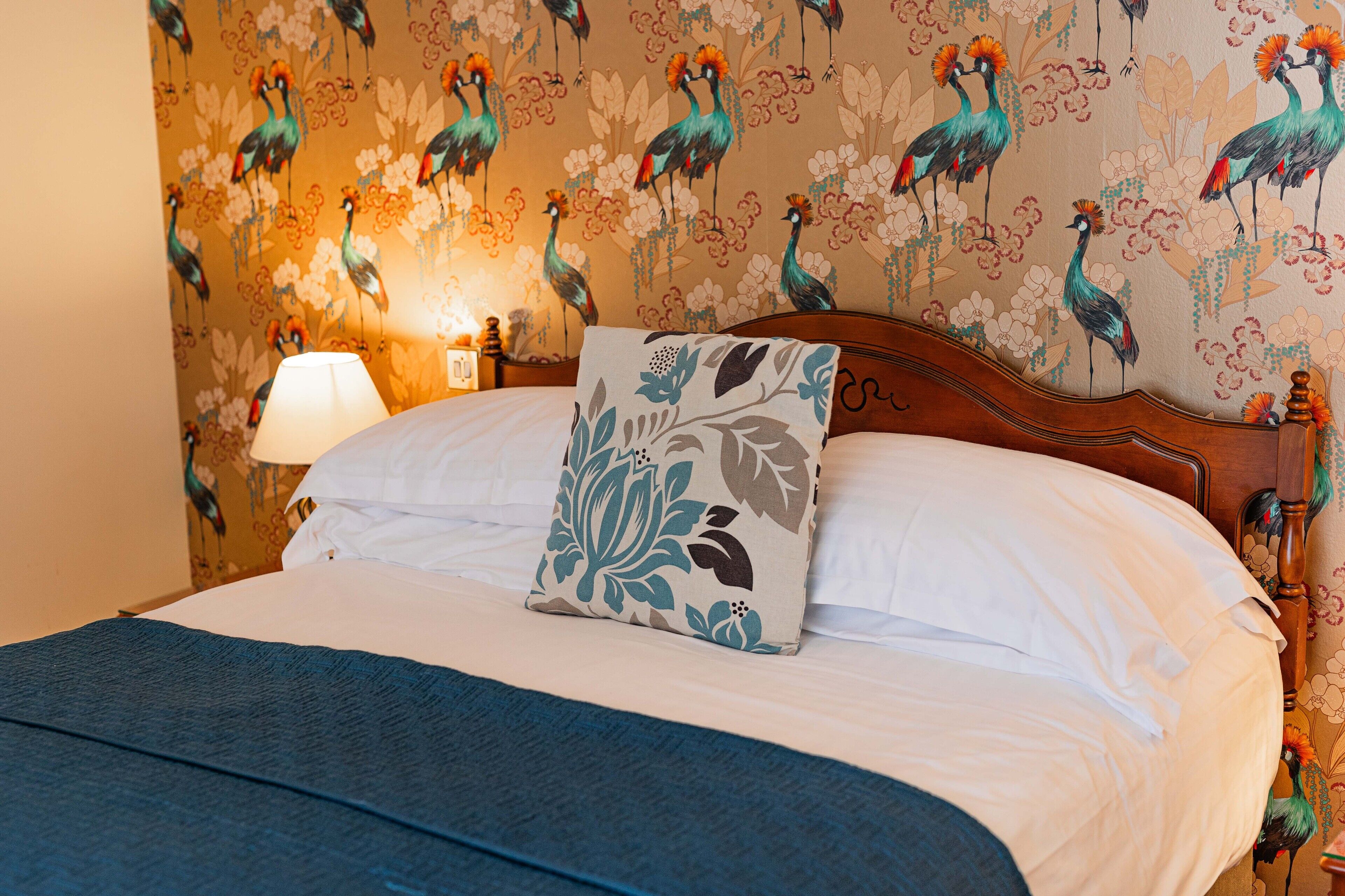 Cama bem arrumada com travesseiro floral, cabeceira de madeira e parede com desenho de pássaros.