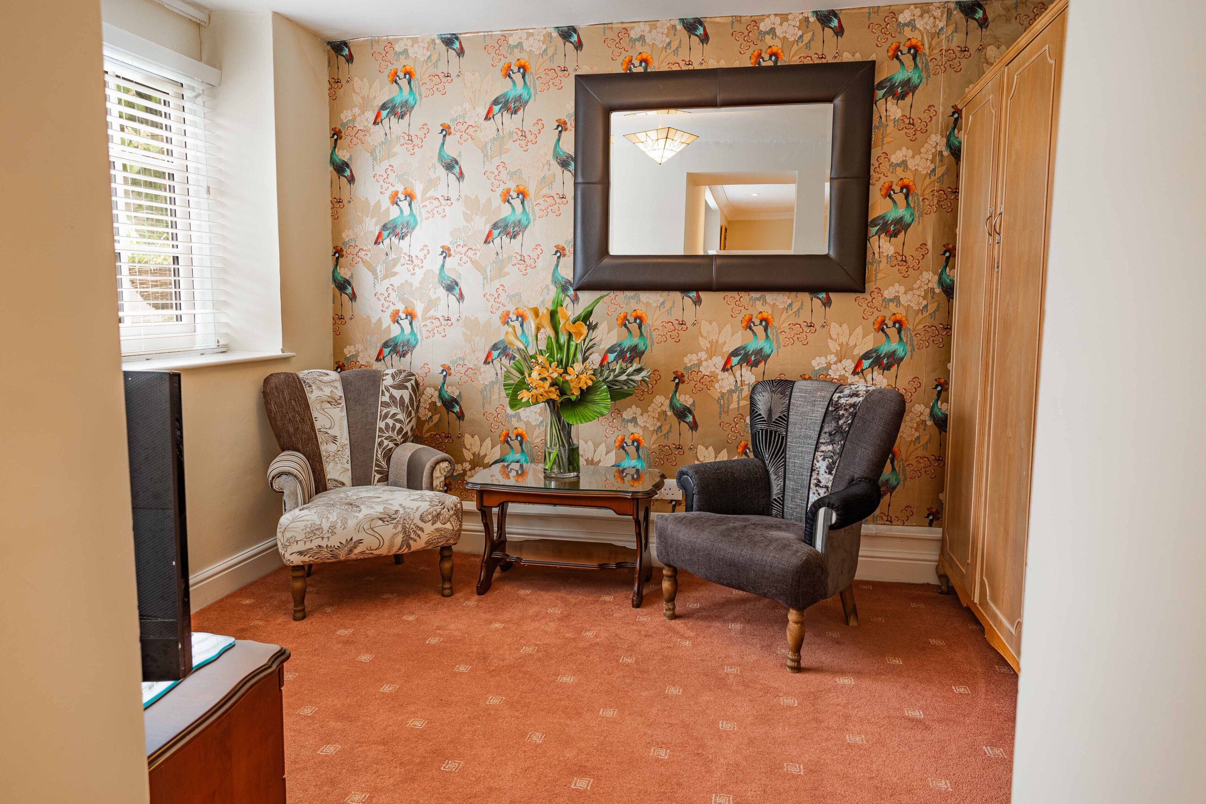 Una habitación con dos sillones estampados, una mesita con un jarrón de flores y un espejo grande en la pared.