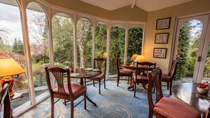 A conservatory with a dining table and chairs, surrounded by large windows offering a view of trees and a garden. - The Burn How Garden House Hotel (Windermere)