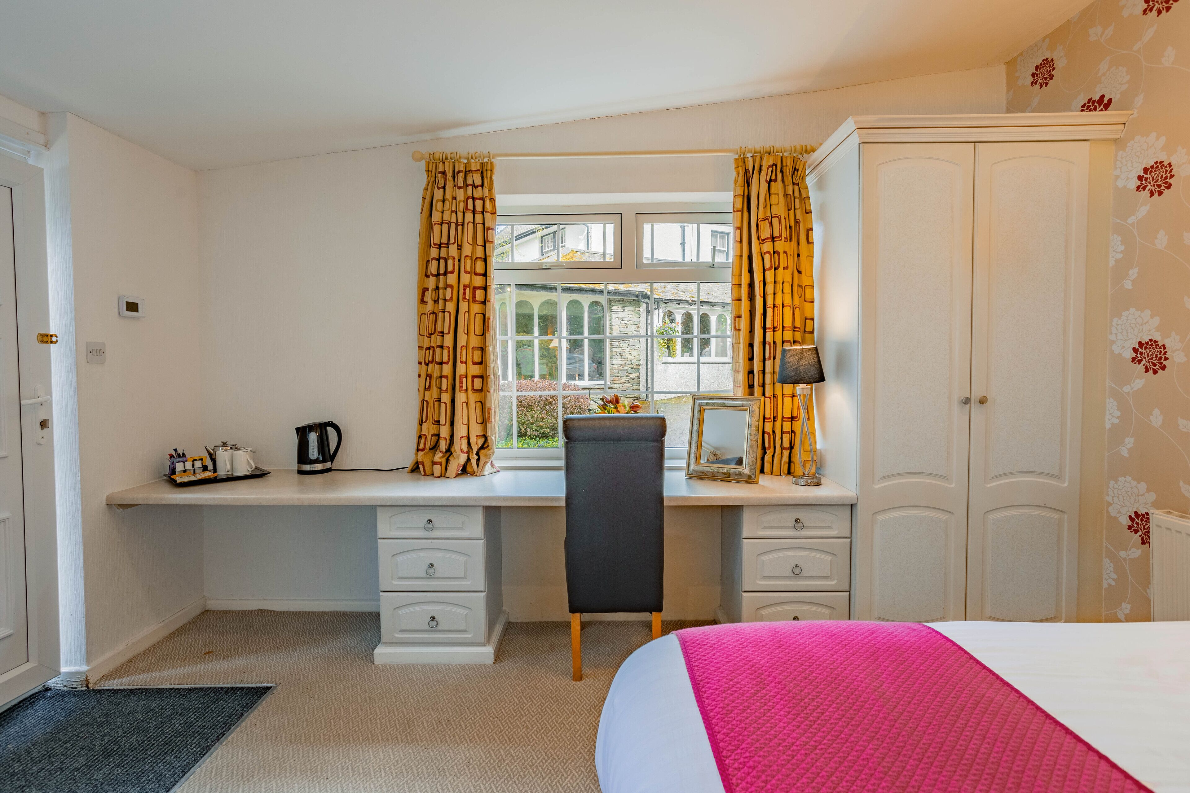 A bedroom with a bed, a desk with a chair, a window with curtains, a wardrobe, and a radiator.