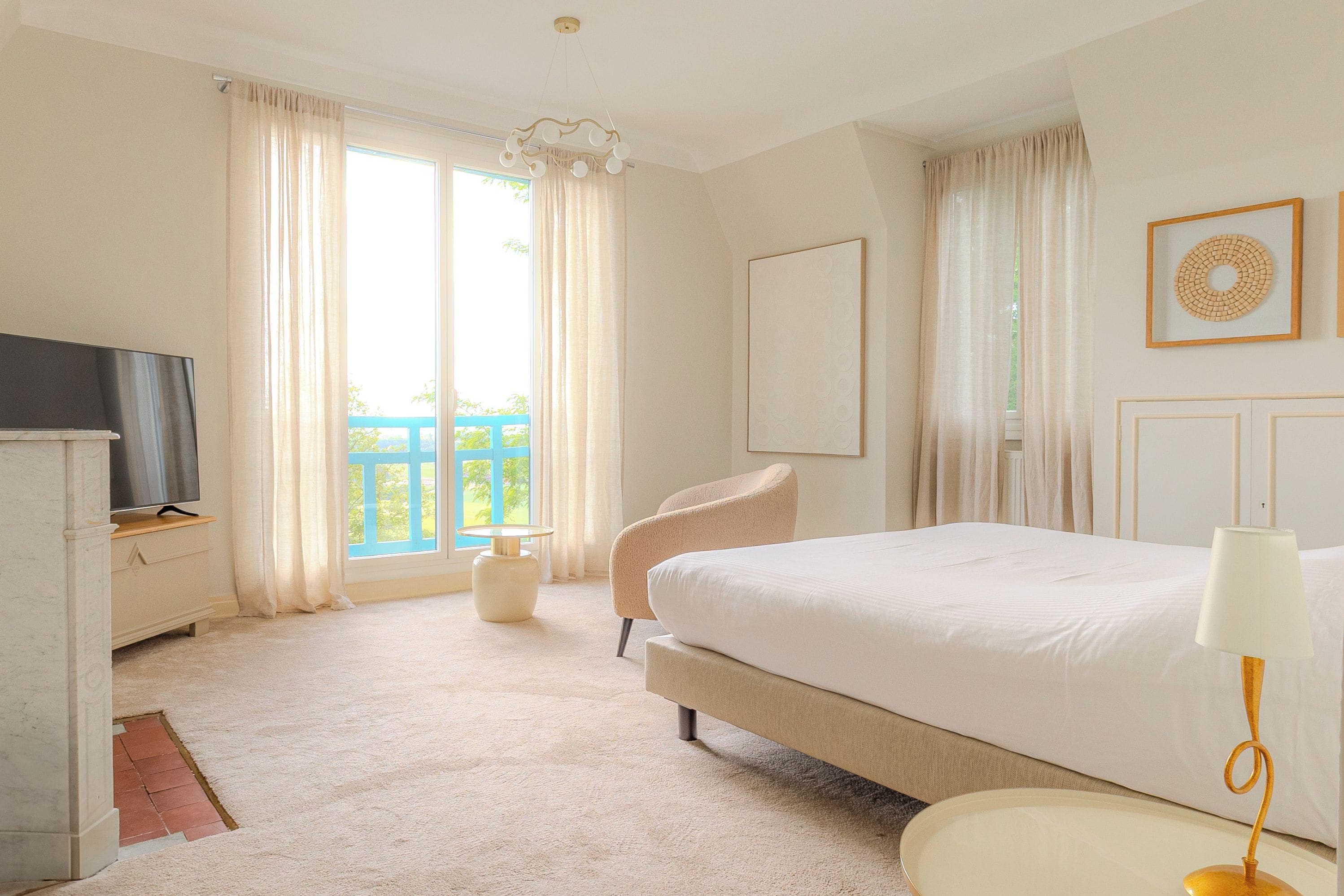 Ein Schlafzimmer mit einem Bett, einem Sessel, einem Fernseher und einem Balkon, der durch das Fenster sichtbar ist.