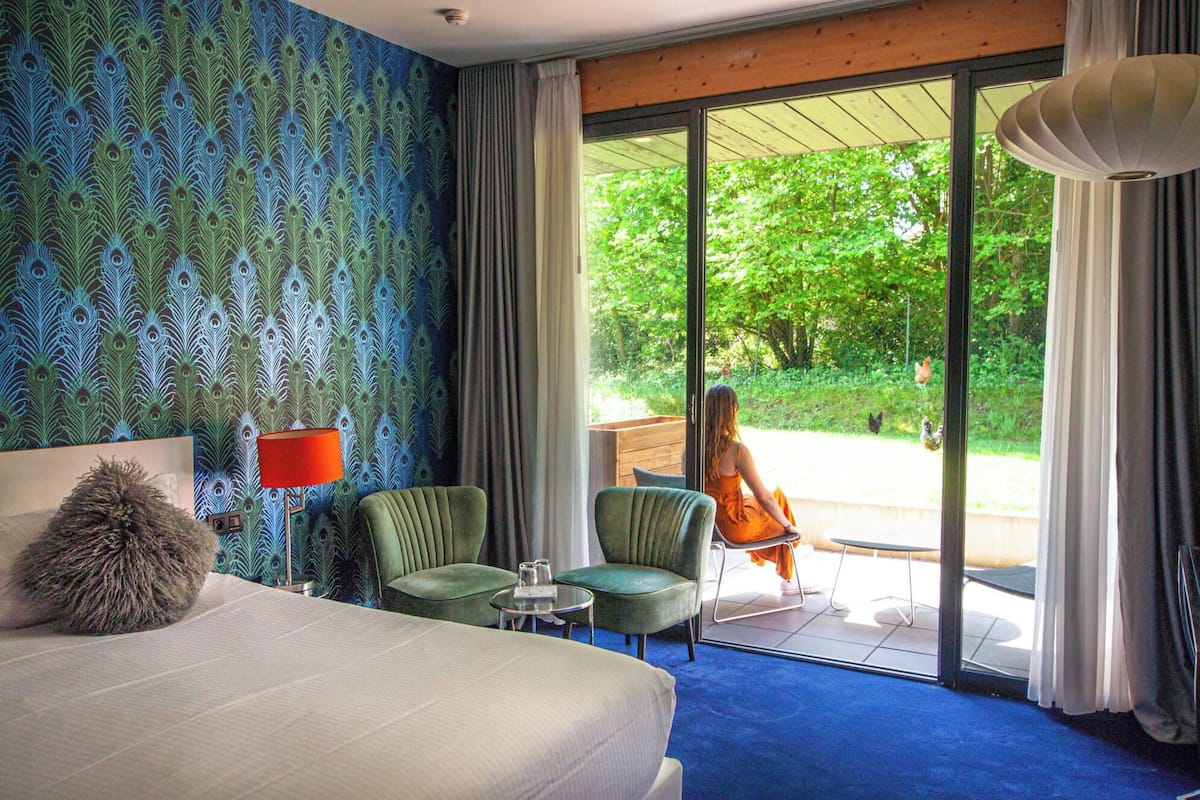Ein Hotelzimmer mit einem großen Bett, zwei Stühlen, einem kleinen Tisch und Blick auf einen Garten durch eine Glastür.