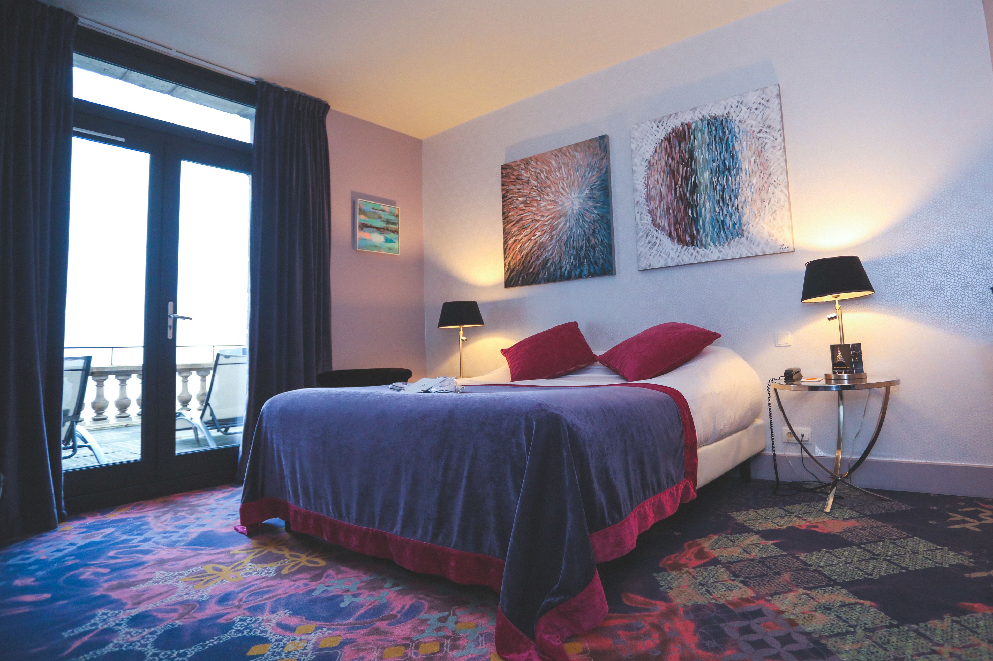 A hotel room with a bed, two side tables with lamps, a balcony view, and artwork on the wall.