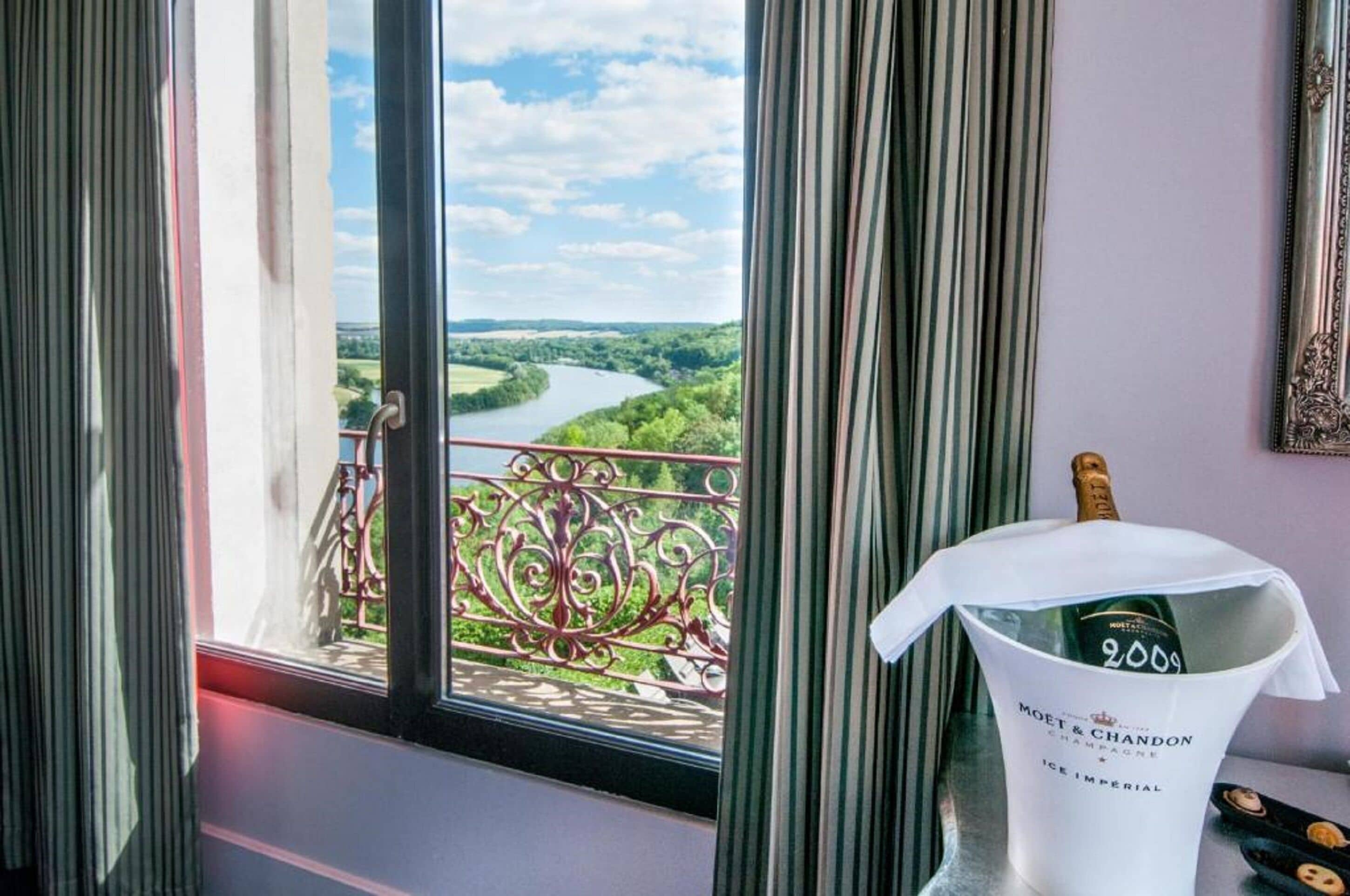Une chambre avec vue sur une rivière et sur la verdure, une bouteille de champagne Moët & Chandon et un garde-corps décoratif en métal.