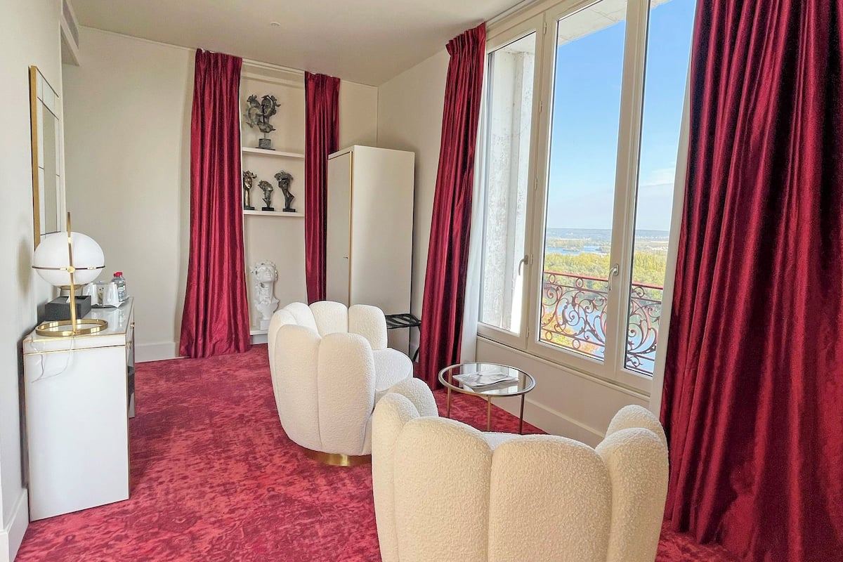 Ein Hotelzimmer mit rotem Teppich, weißen Sesseln, einem kleinen Tisch, einer Lampe, einem Schminktisch und Blick ins Freie durch große Fenster.
