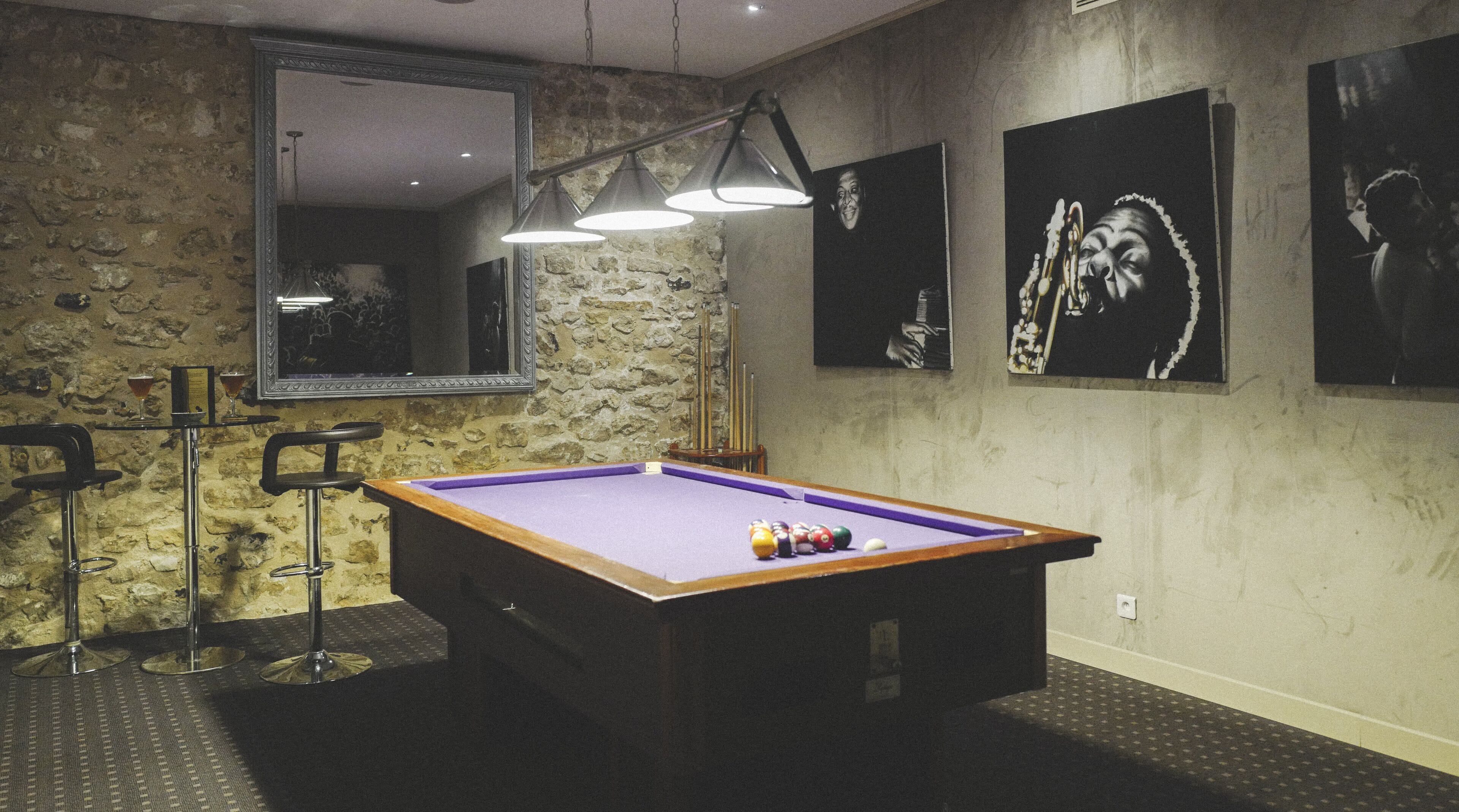 A pool table with balls, a mirror, and pool cues in a room with stone walls and artwork.