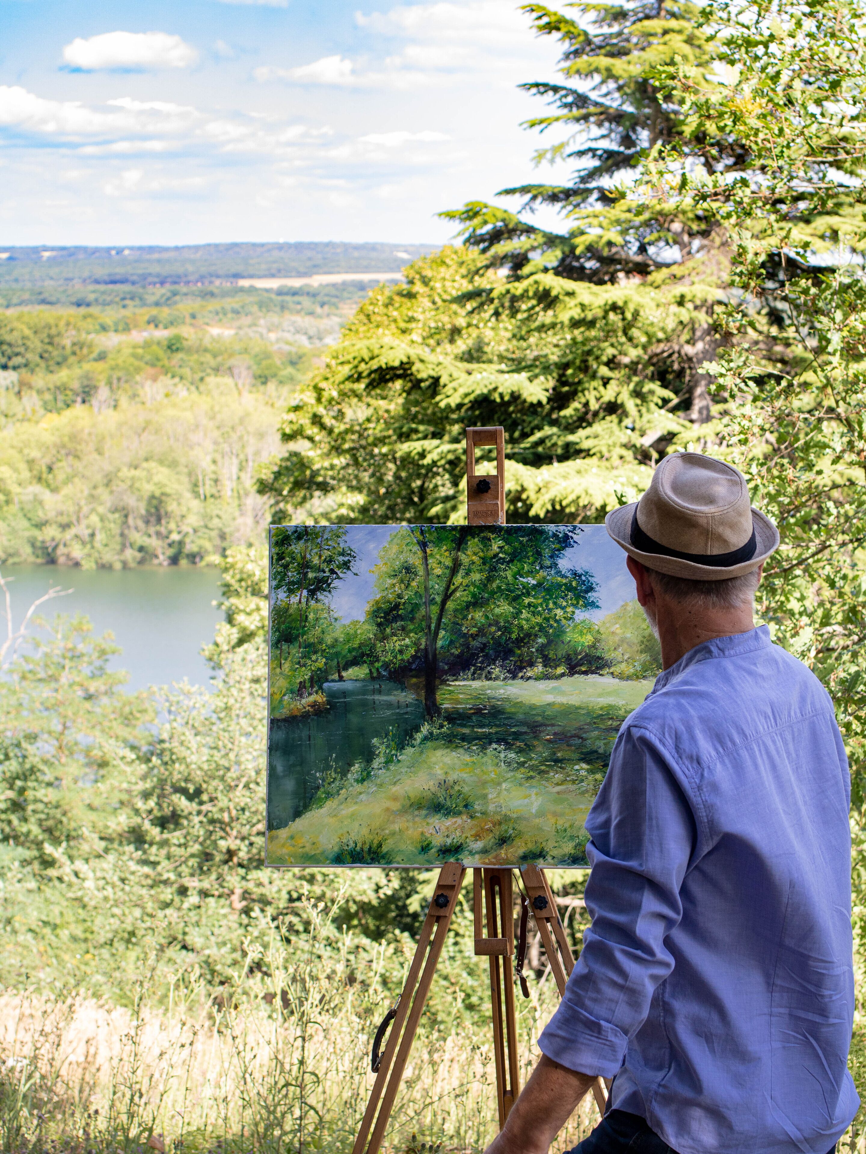 A man painting a landscape on an easel with a view of a lake and trees in the background.