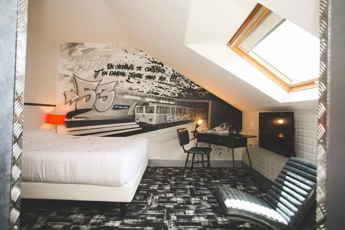 Ein kompakter Raum mit Bett, Schreibtisch und Stuhl. An der Wand ist ein Wandbild mit einem Zug und Text. Über dem Schreibtisch befindet sich ein Oberlicht.