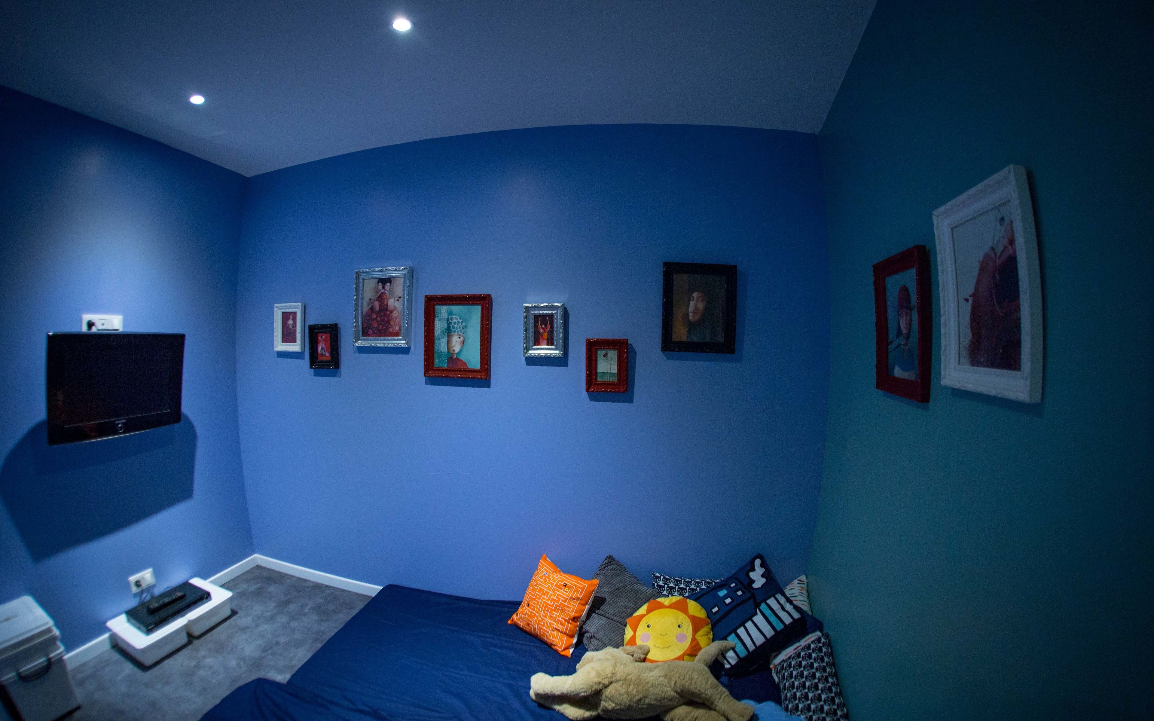 A bedroom with a blue wall, a bed with multiple pillows, a flat-screen TV mounted on the wall, and several framed pictures.