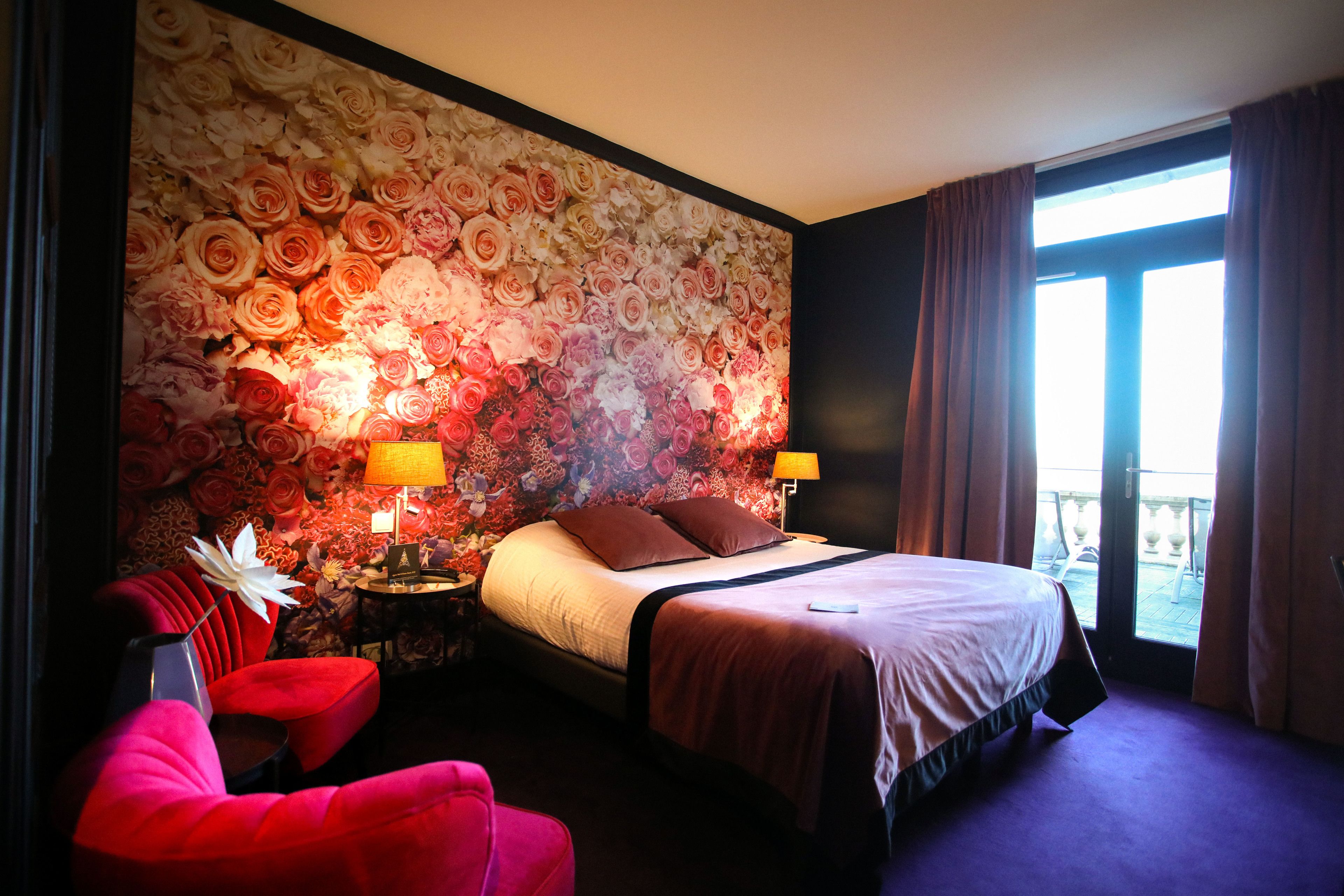 Habitación de hotel con un amplio papel tapiz floral, una cama con cabecera marrón, dos lámparas de noche, un sillón rojo y vistas al exterior a través de un gran ventanal.