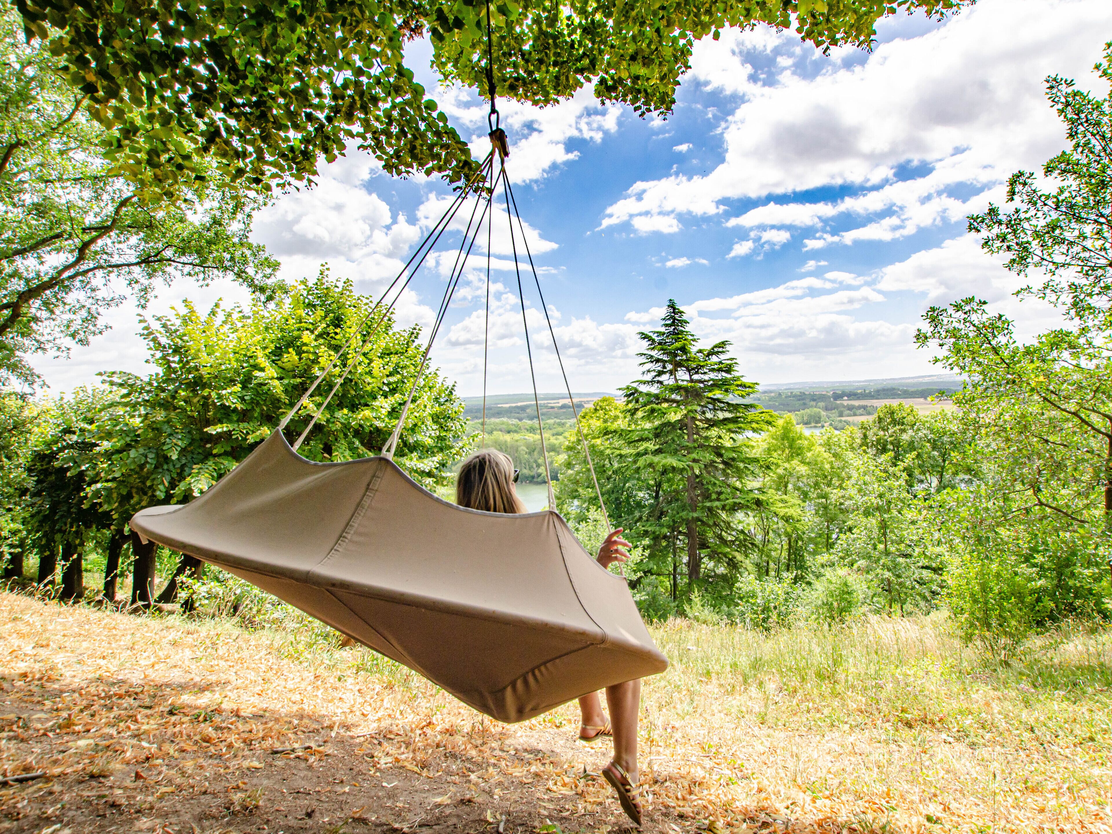 A person on a hammock swing in a natural setting with trees and a view of the landscape.