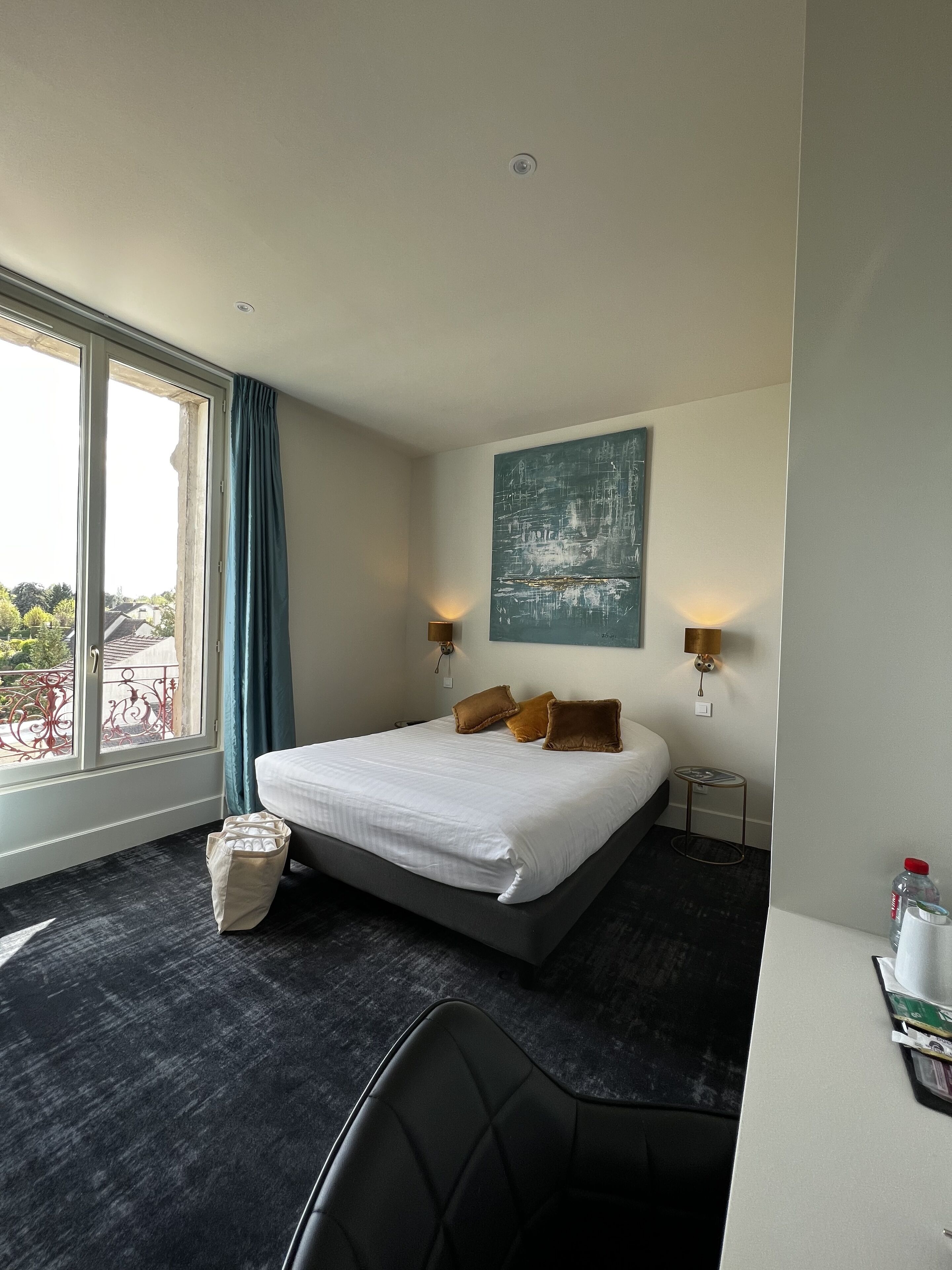 Habitación de hotel con una cama grande, dos mesitas de noche con lámparas, una mesita auxiliar pequeña, una silla, un ventanal con cortinas y vistas al exterior.
