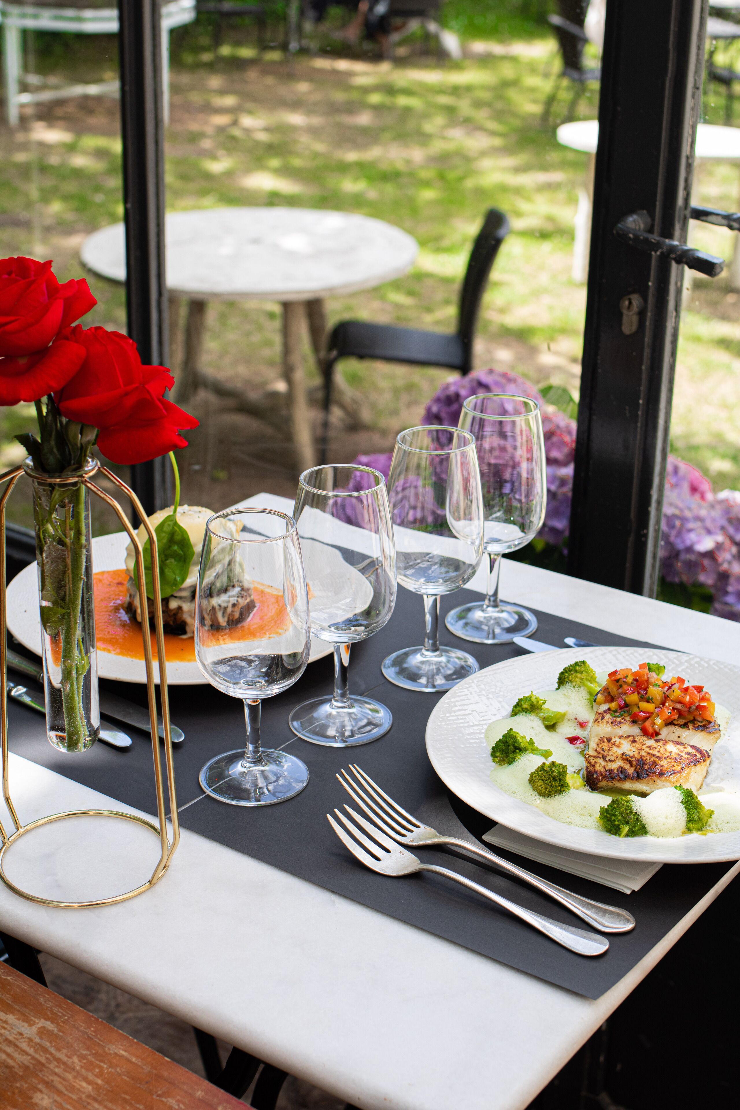Une table à manger dressée avec des assiettes, des verres à vin et des couverts, agrémentée d’un vase de roses rouges et offrant une vue sur le jardin.