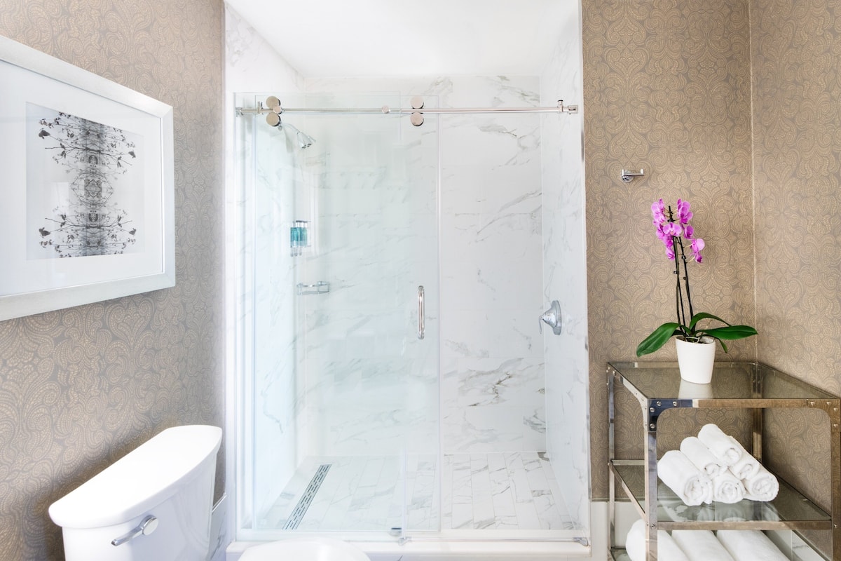 A modern bathroom with a glass-enclosed shower, a toilet, a framed artwork, and a potted orchid.