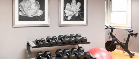 Een sportschool met trainingsapparatuur, waaronder gewichten, een rode fitnessbal en een hometrainer. Er hangen twee ingelijste foto's aan de muur.