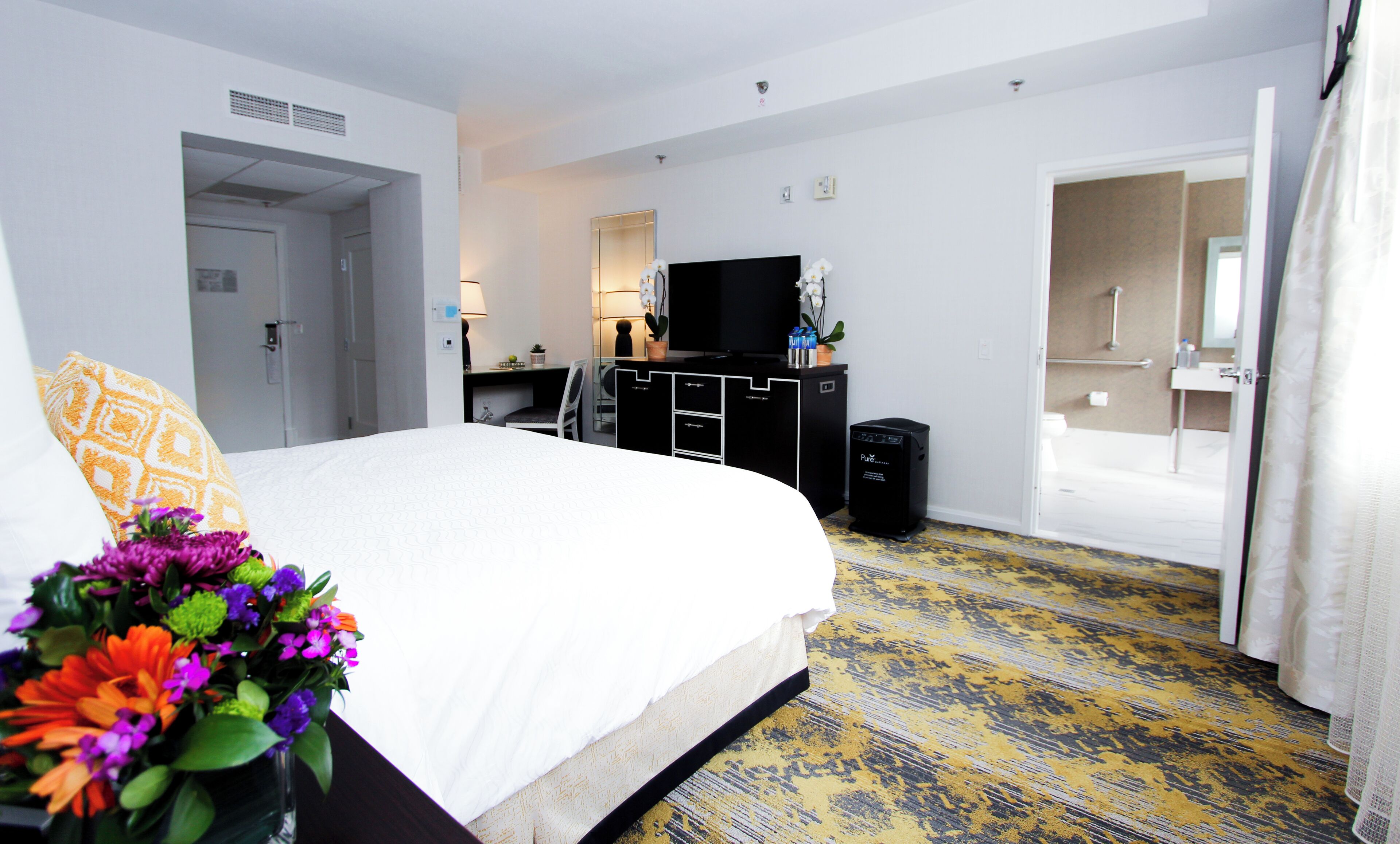 A hotel room with a bed, a desk, a TV, a trash bin, and a bathroom visible through an open door.