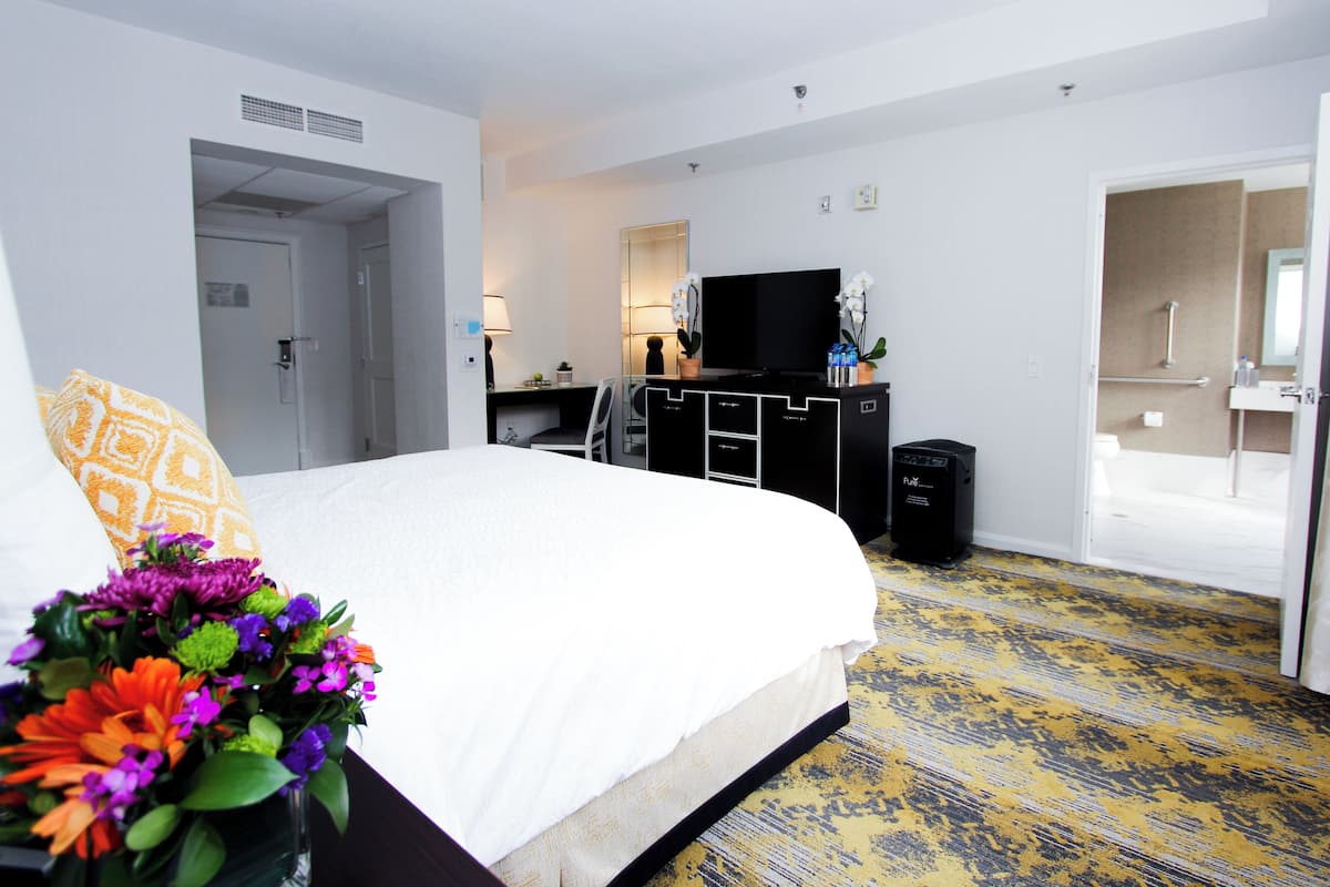 A hotel room with a bed, a desk, a TV, a trash bin, and a bathroom visible through an open door.
