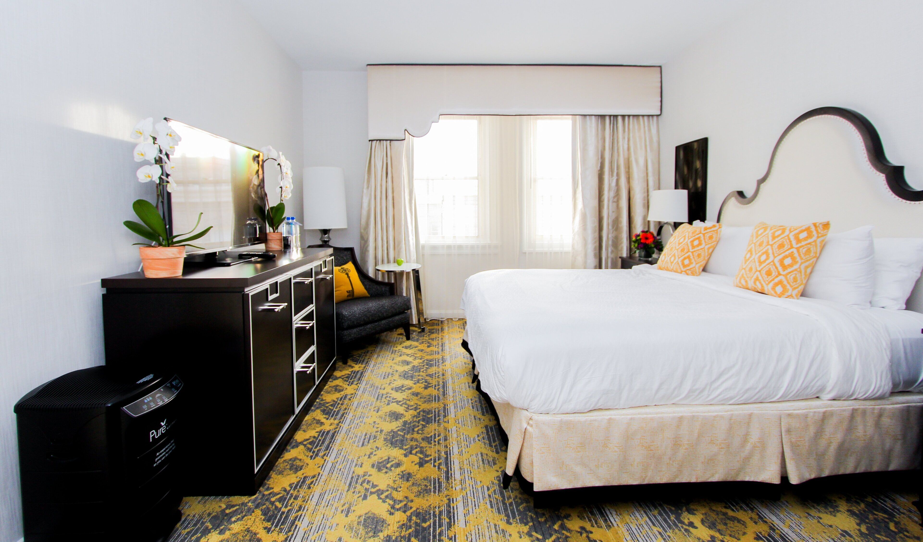 A hotel room with a bed, a desk, a chair, a dresser, and a window with curtains.