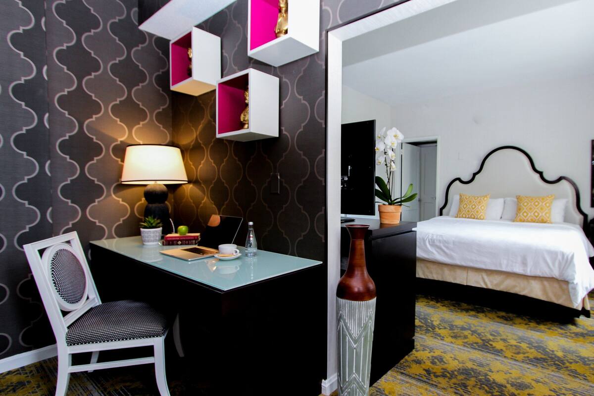 A hotel room with a bed, a desk, a chair, a lamp, and a potted plant.