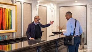 A man in a suit gesturing to another man in a blue shirt standing at a reception desk in a hotel lobby with a framed artwork on the wall. - The Churchill Hotel near Embassy Row (Washington)