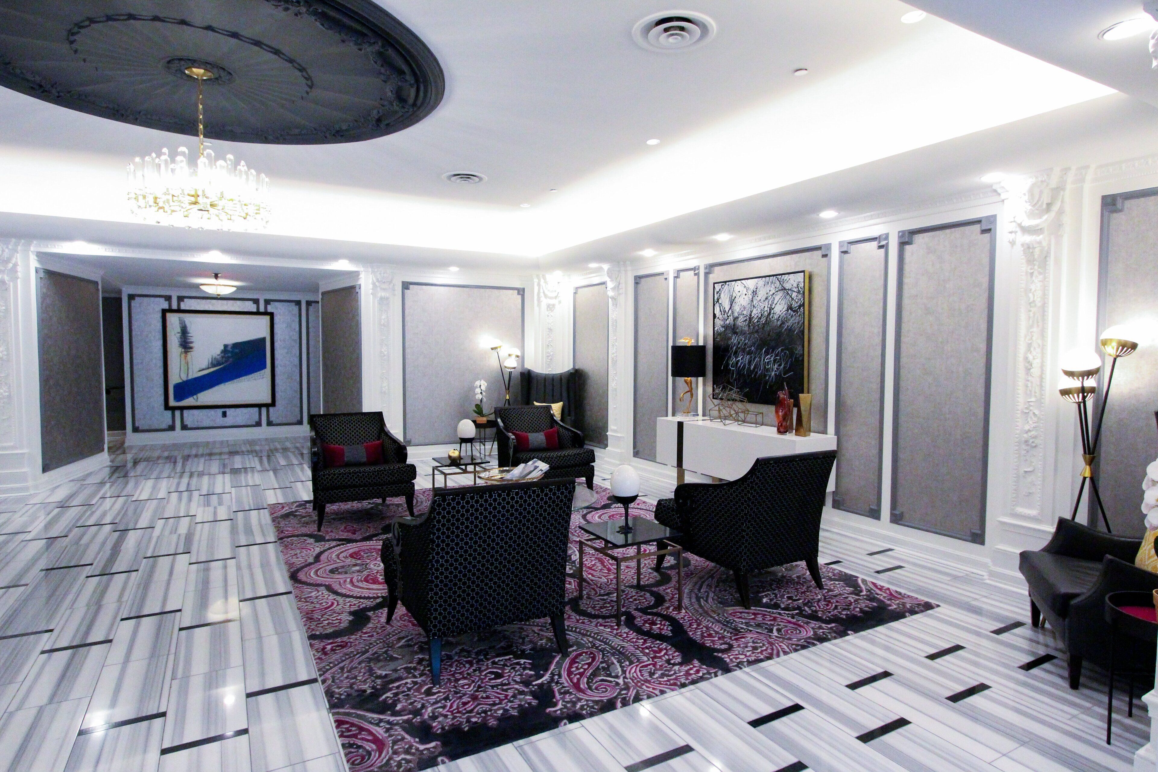 Een moderne hotelentree met een marmeren vloer, een kroonluchter, een receptiebalie en een zithoek met zwarte stoelen en een vloerkleed met dessin.