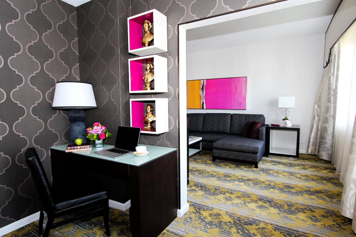 A modern hotel room with a desk, chair, and a sofa. There are decorative items on the desk and a lamp. The room has patterned wallpaper and a carpet with a geometric design.