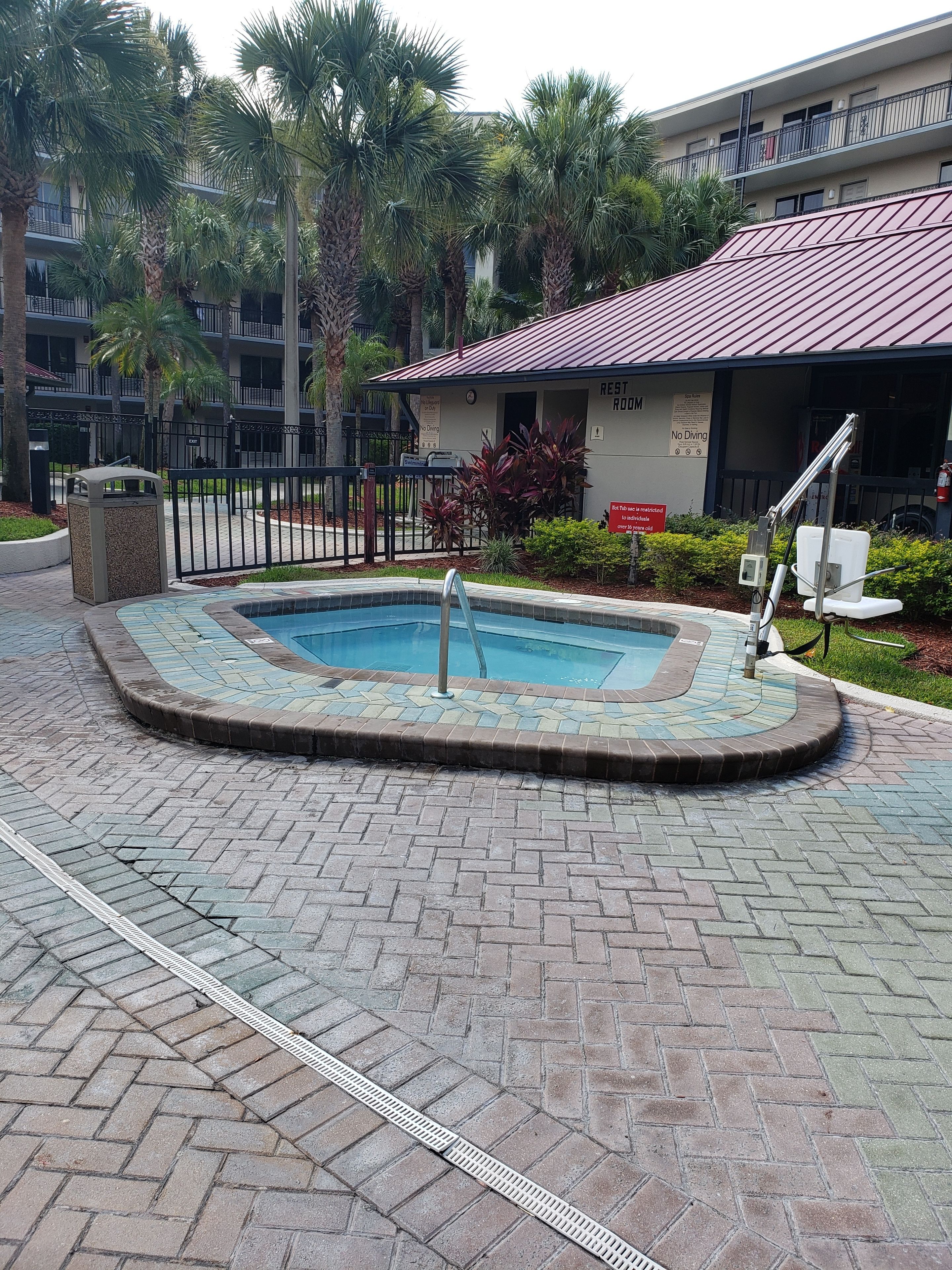 A pool area with a brick pathway, a metal fence, and a building with a red roof labeled "REST ROOM".