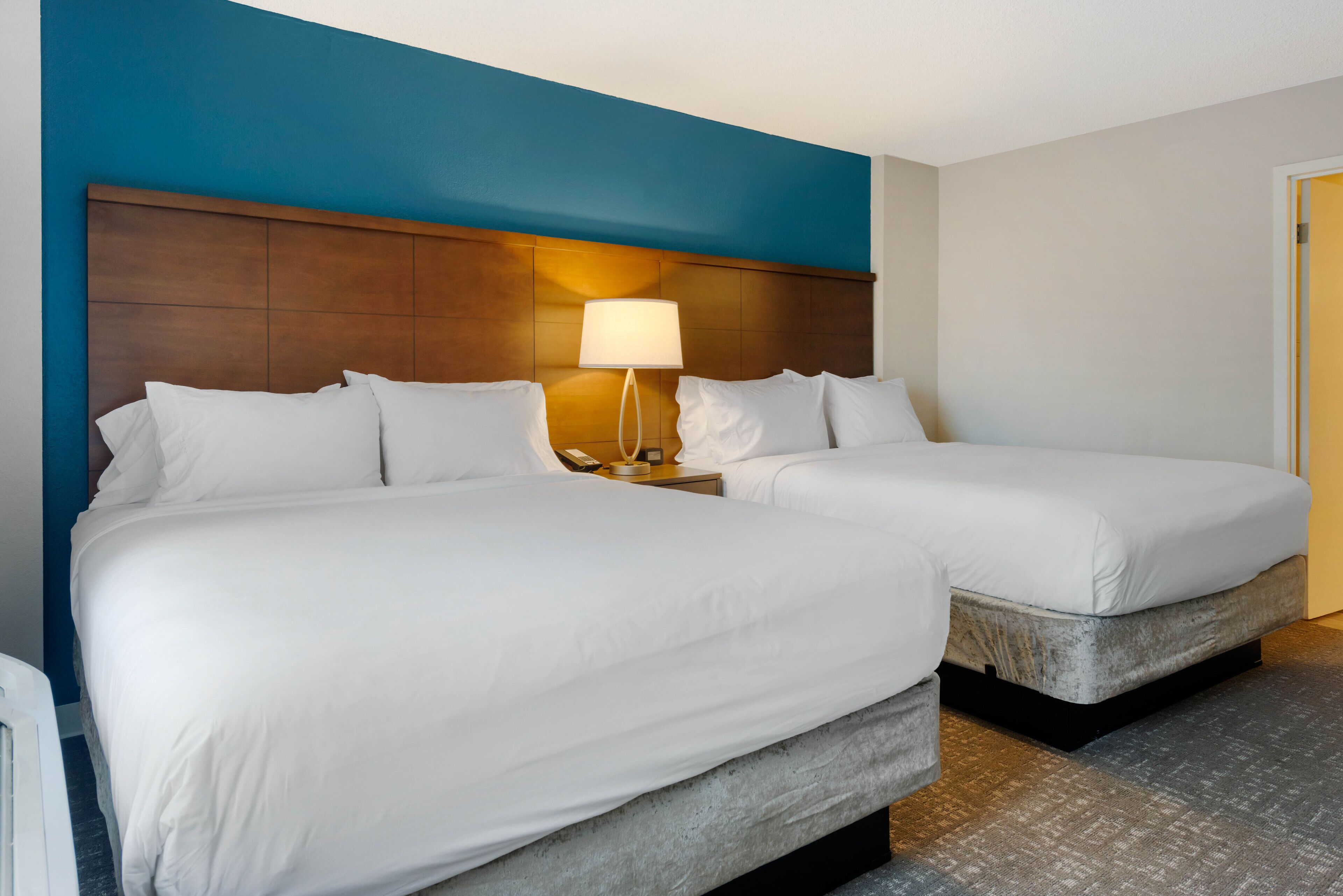 Habitación de hotel con dos camas, una mesita de noche con lámpara y una pared azul decorativa.