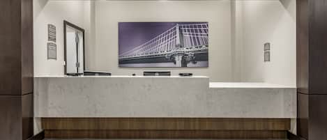 A reception desk with a 'Please Wait Here' sign, a framed picture of a bridge, and a wooden panel wall.