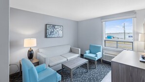 Une chambre d’hôtel avec un canapé blanc, deux fauteuils bleus, une petite table en bois, un bureau avec une lampe et une grande fenêtre donnant sur l’extérieur.