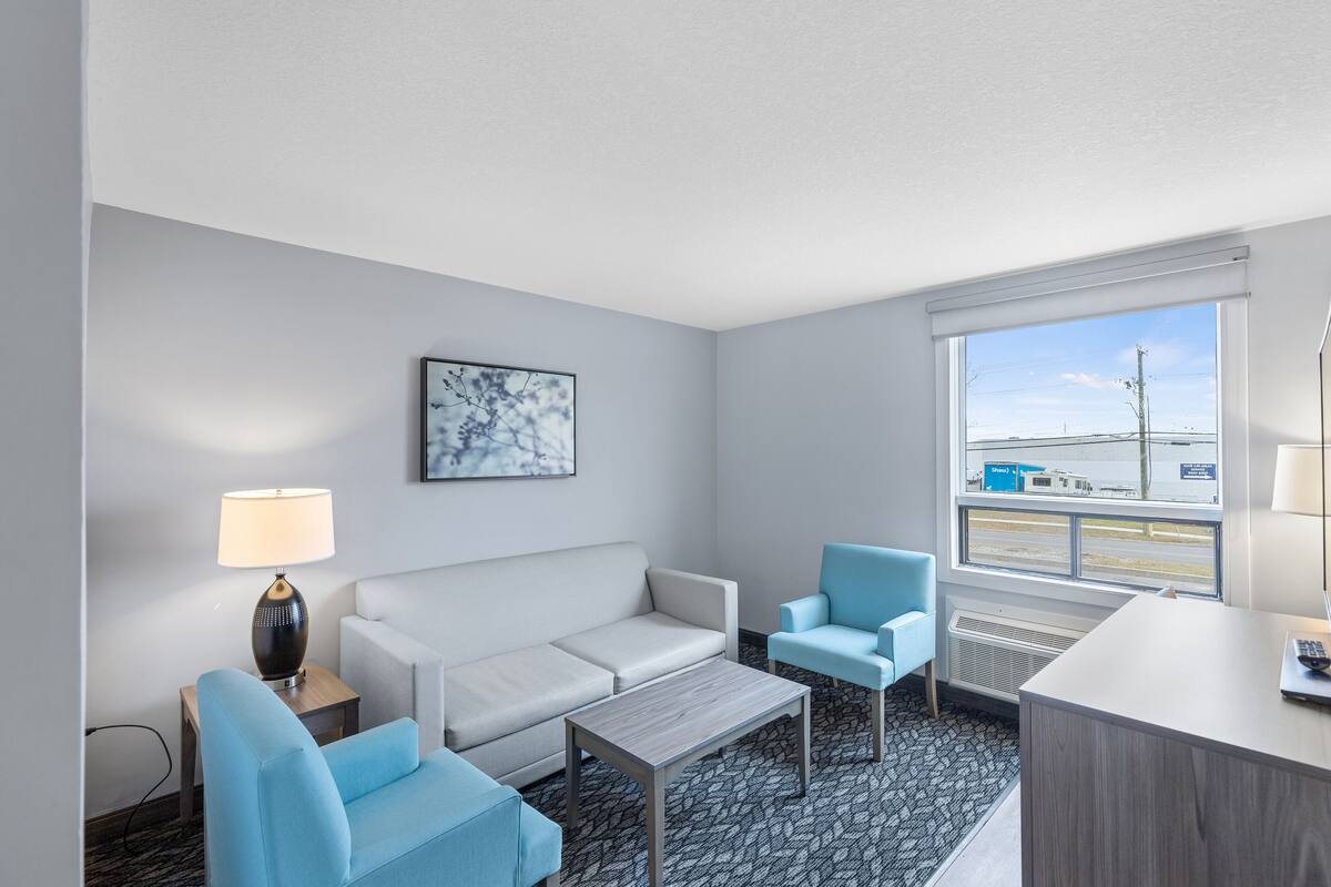Une chambre d’hôtel avec un canapé blanc, deux fauteuils bleus, une petite table en bois, un bureau avec une lampe et une grande fenêtre donnant sur l’extérieur.