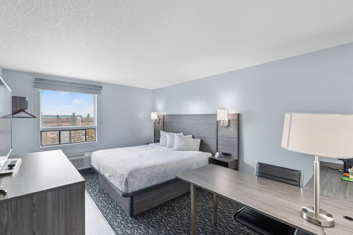 Une chambre d’hôtel moderne, équipée d’un lit, d’un bureau et d’une grande fenêtre donnant sur le paysage urbain.