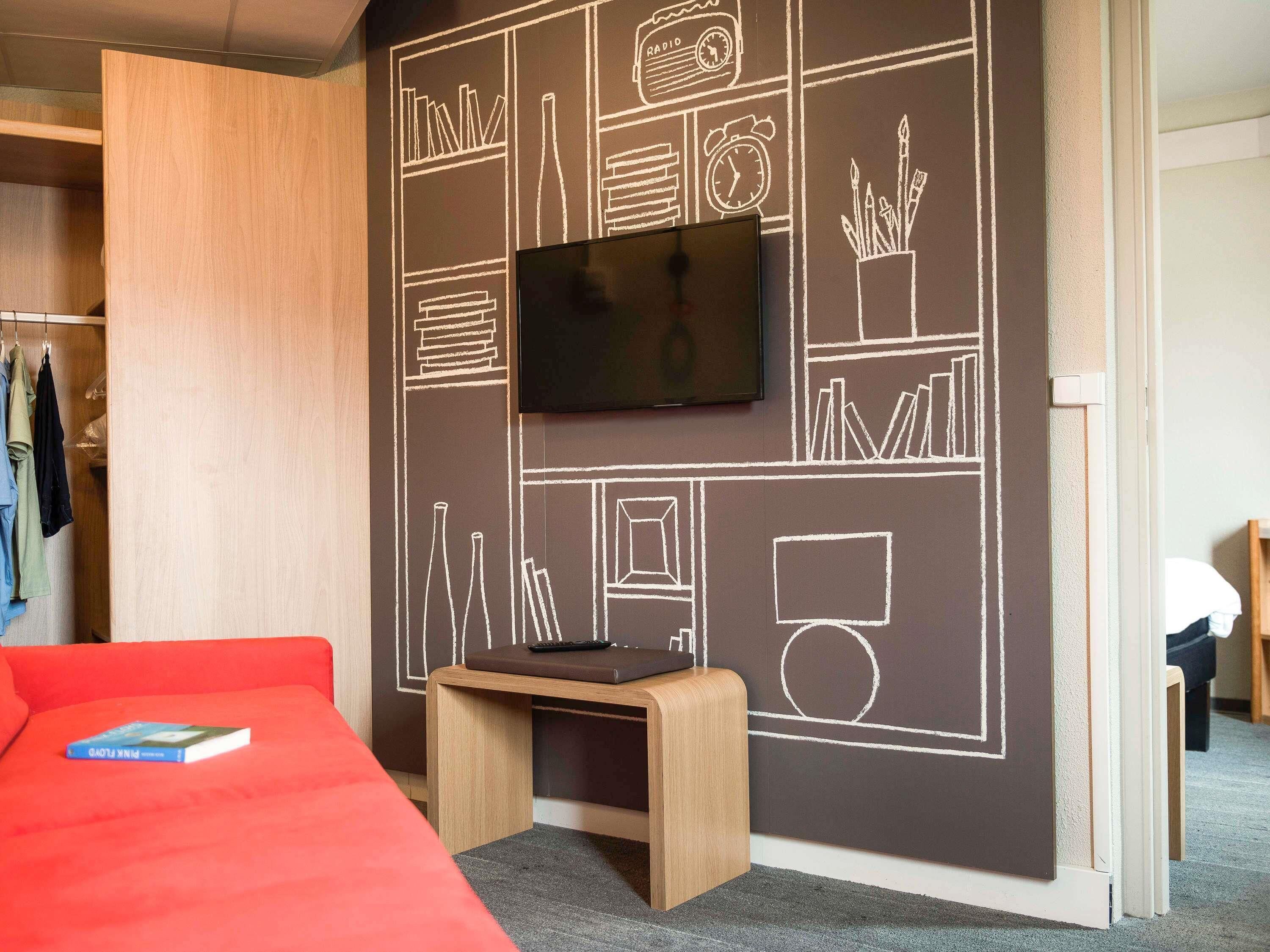 A modern room with a large flat-screen TV mounted on a chalkboard wall, a wooden wardrobe, a red sofa, and a small wooden table.
