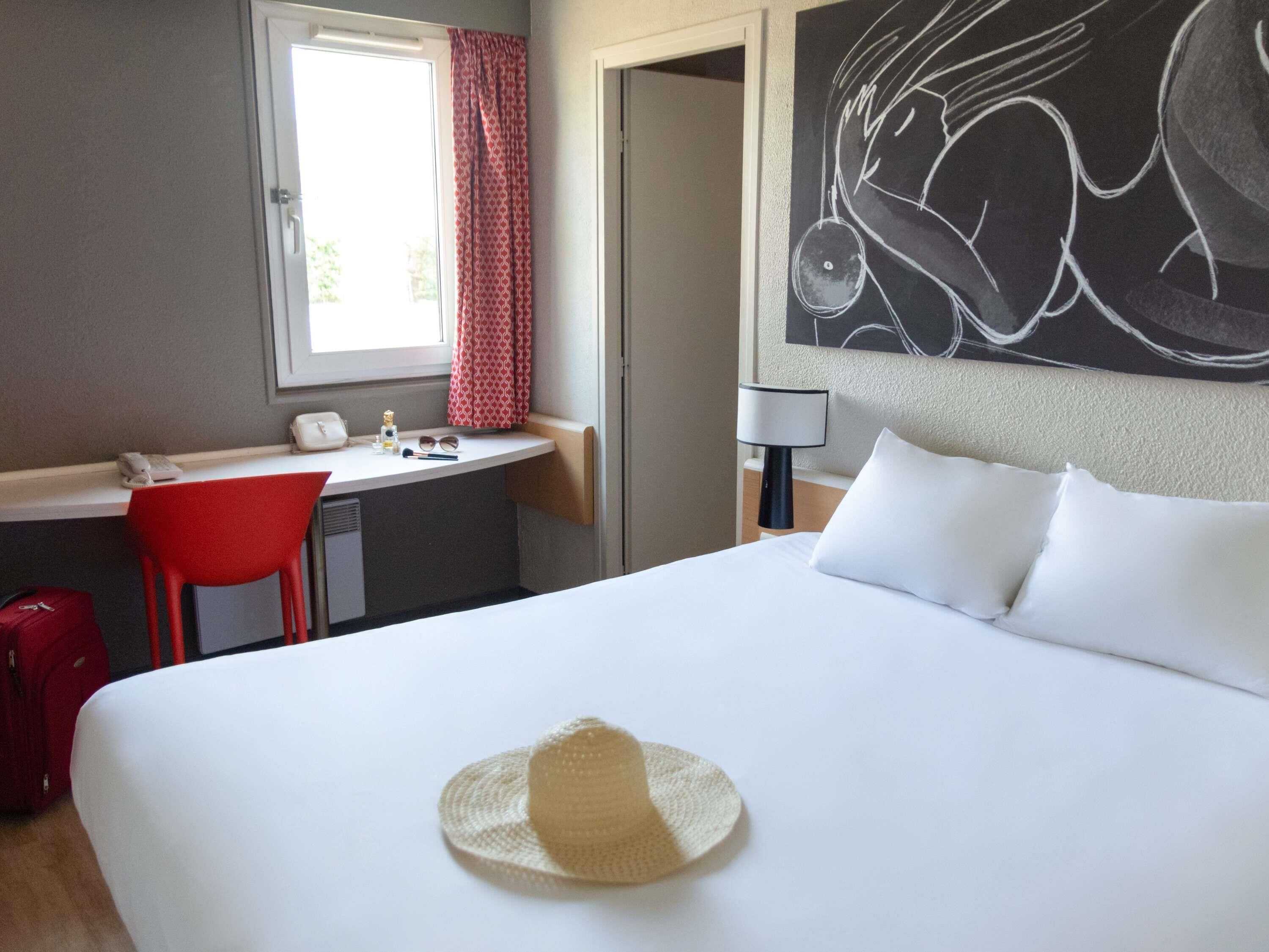 Ein Hotelzimmer mit einem Bett, einem Schreibtisch, einem Stuhl, einem Fenster mit Vorhängen, einem Wandbild und einem Hut auf dem Bett.