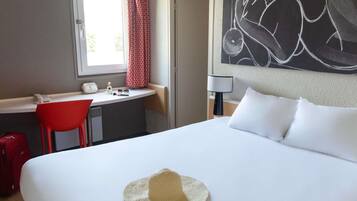 Ein Hotelzimmer mit einem Bett, einem Schreibtisch, einem Stuhl, einem Fenster mit Vorhängen, einem Wandbild und einem Hut auf dem Bett.