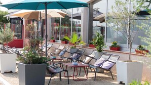 Un espace extérieur aménagé avec des chaises et des tables, des parasols, des plantes en pot et un bâtiment doté de grandes fenêtres.