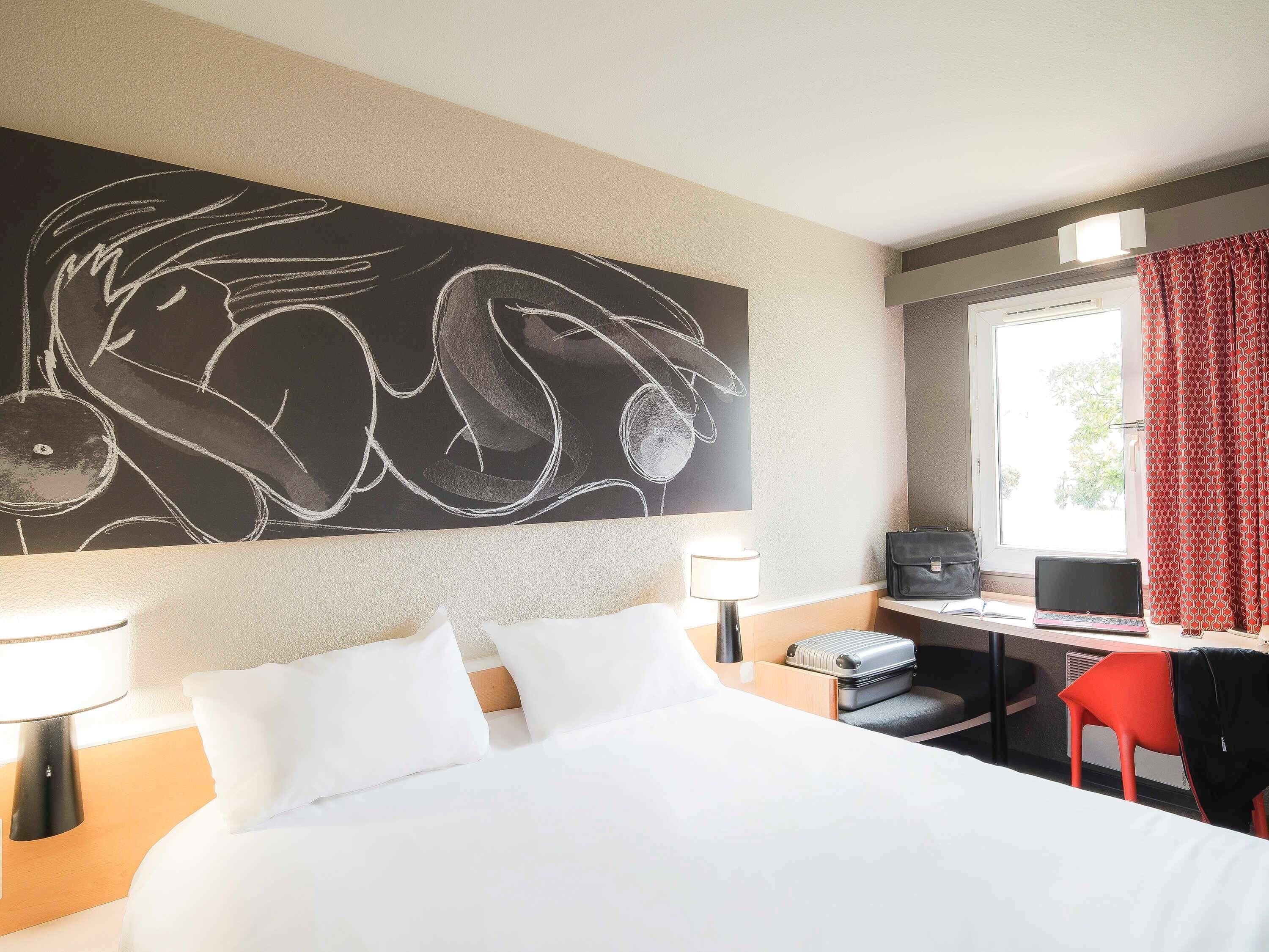 A hotel room with a bed, desk, chair, and a large abstract painting on the wall.
