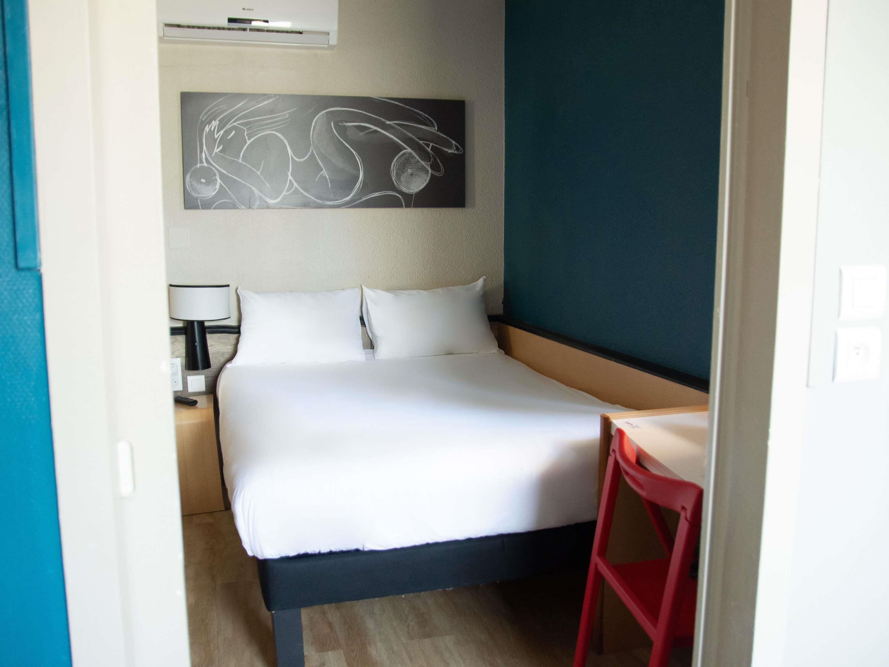 Une chambre d’hôtel équipée d’un lit, d’un bureau, d’une chaise, d’une lampe et d’un climatiseur fixé au mur.