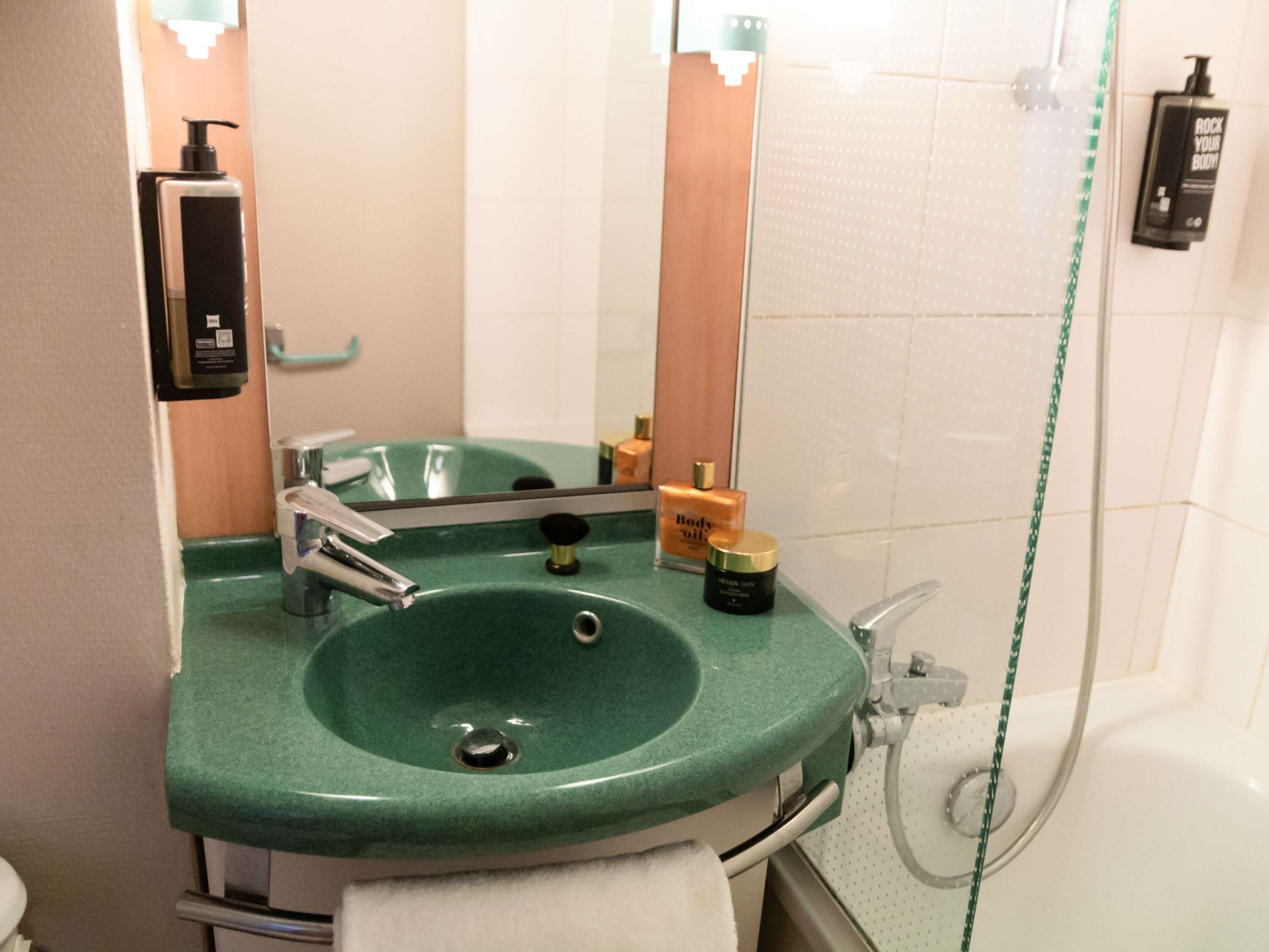 Une salle de bain avec un lavabo vert, un miroir, une douchette à main et une bouteille d’huile pour le corps.